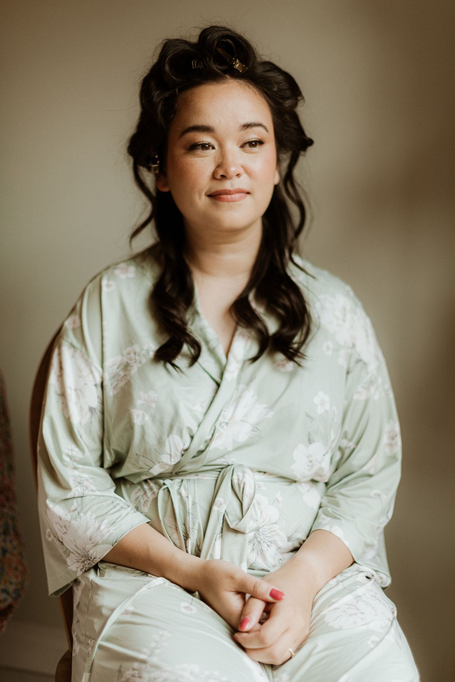 Bride in sage green floral robe sitting and smiling during wedding preparations.