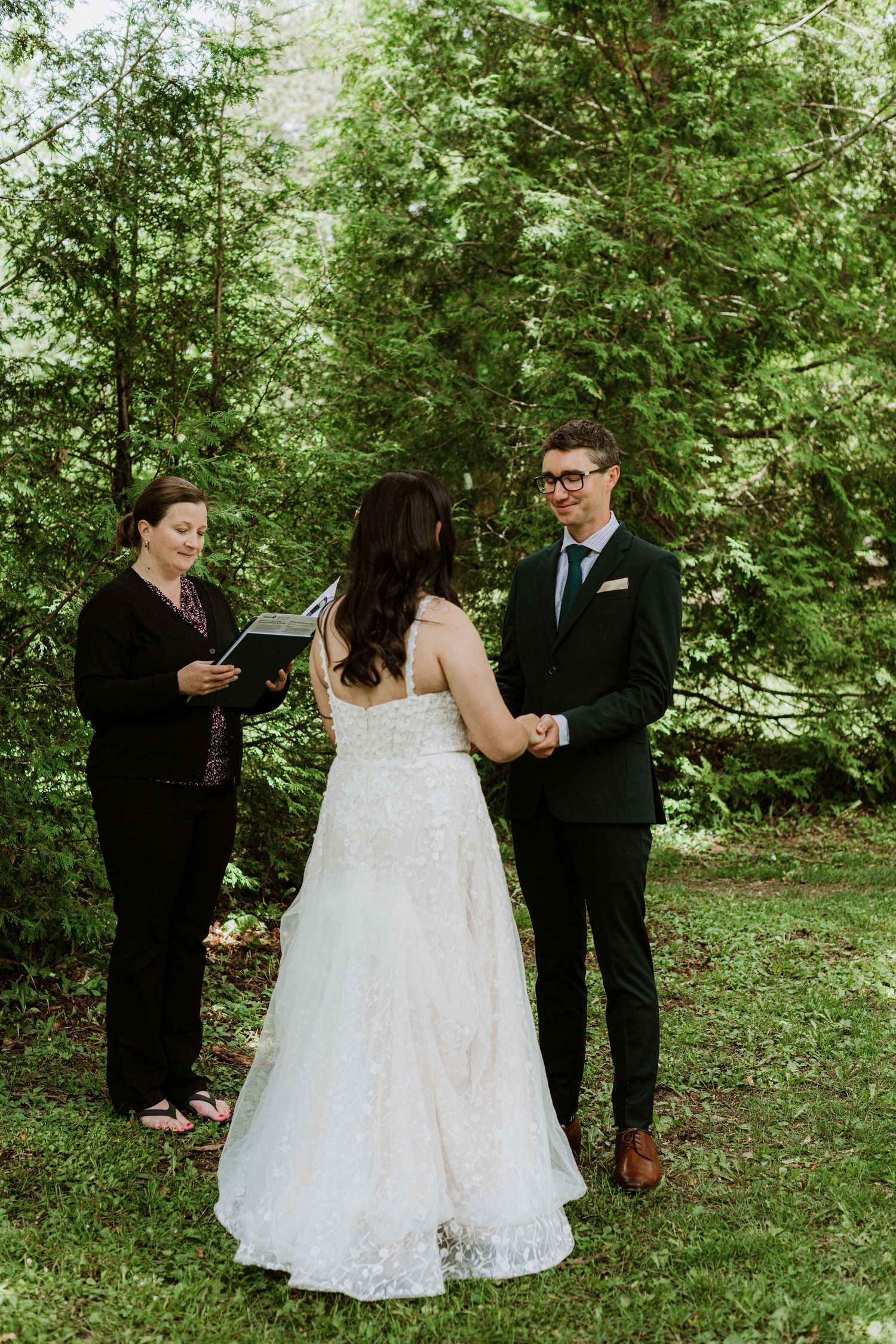 Officiant reads vows as bride and groom hold hands during intimate outdoor forest wedding ceremony.