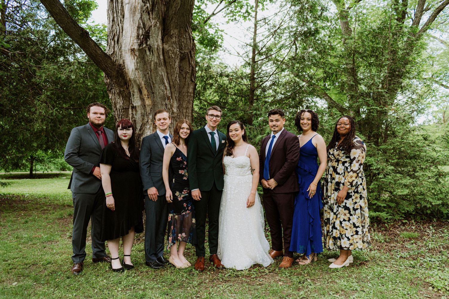 Wedding party group portrait of eight people posing in front of large tree in park.