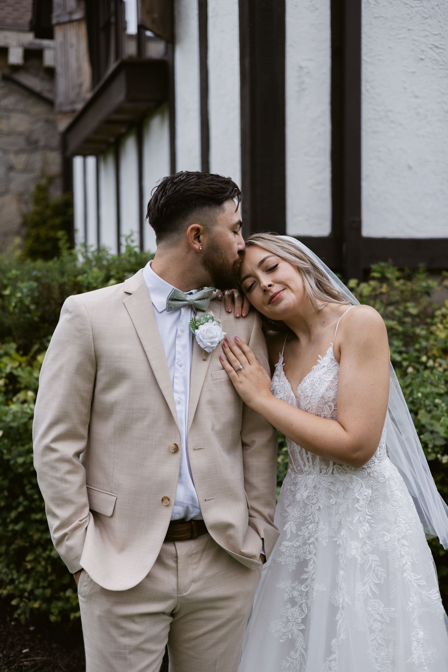 Groom kissing bride on the cheek outdoors, both in elegant wedding attire with Tudor-style building behind.