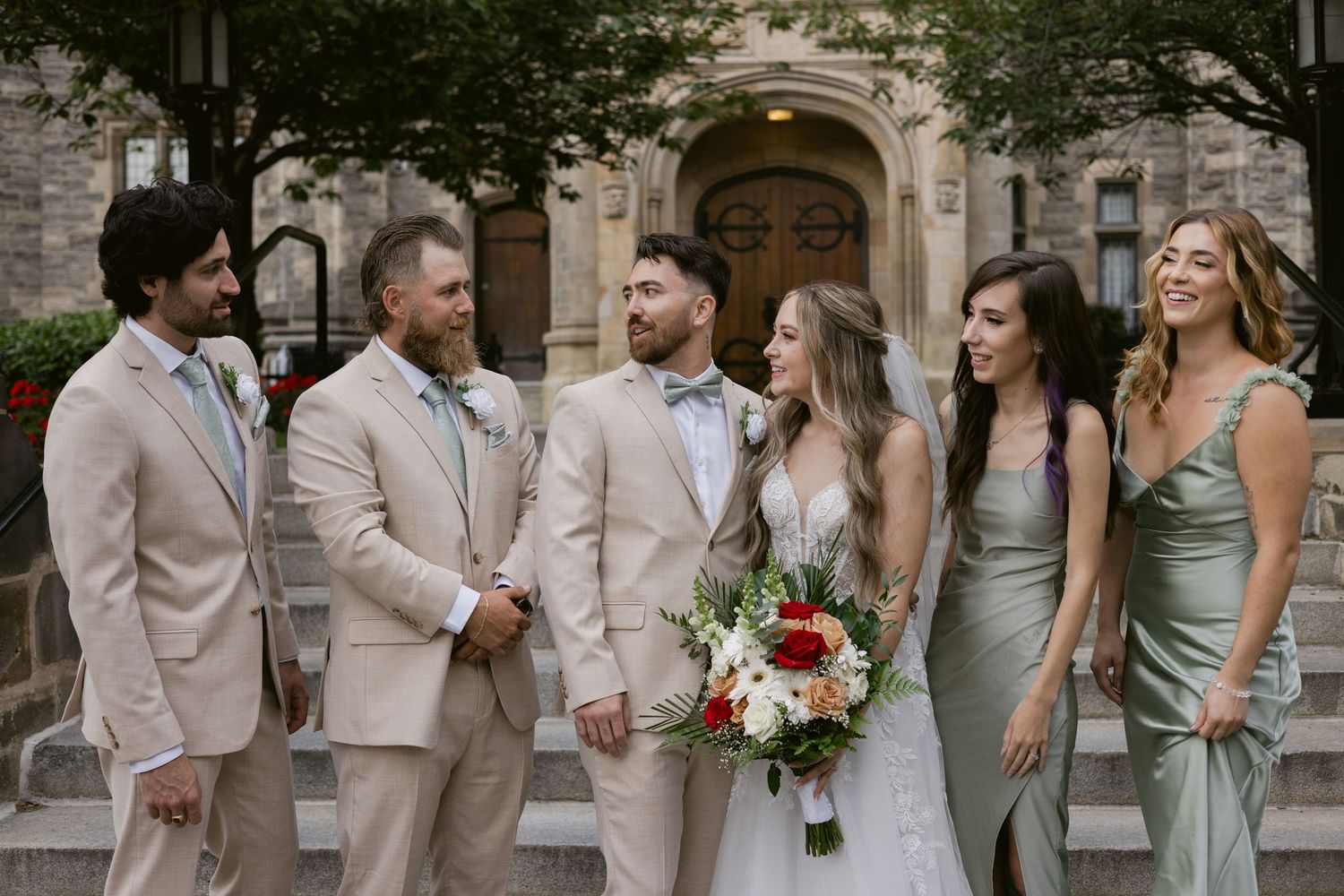 Smiling wedding party laughs together on stone steps, bridesmaids in sage satin gowns and groomsmen in tan suits.