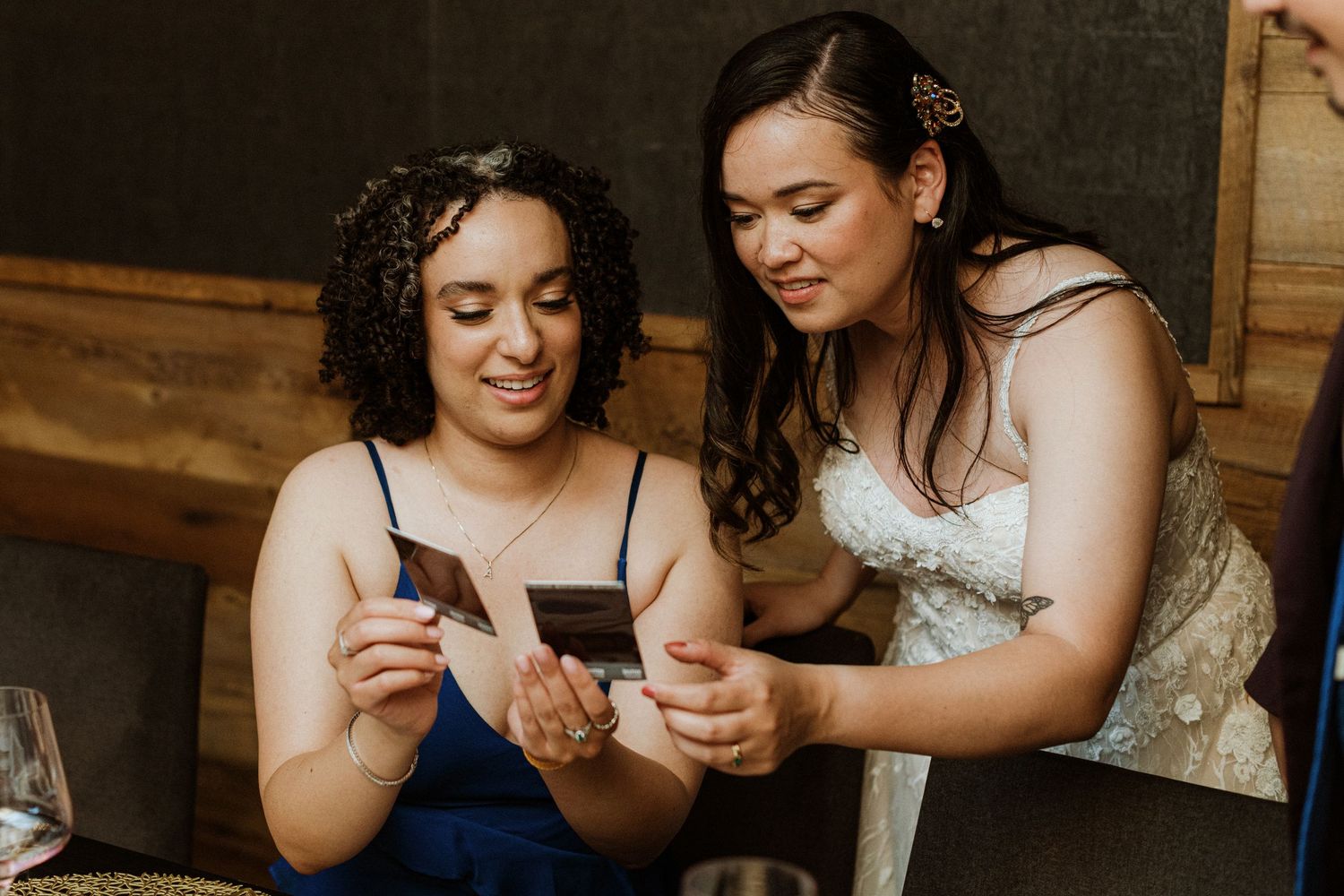 Bride in white gown shares Polaroid photos with guest in blue dress at reception.