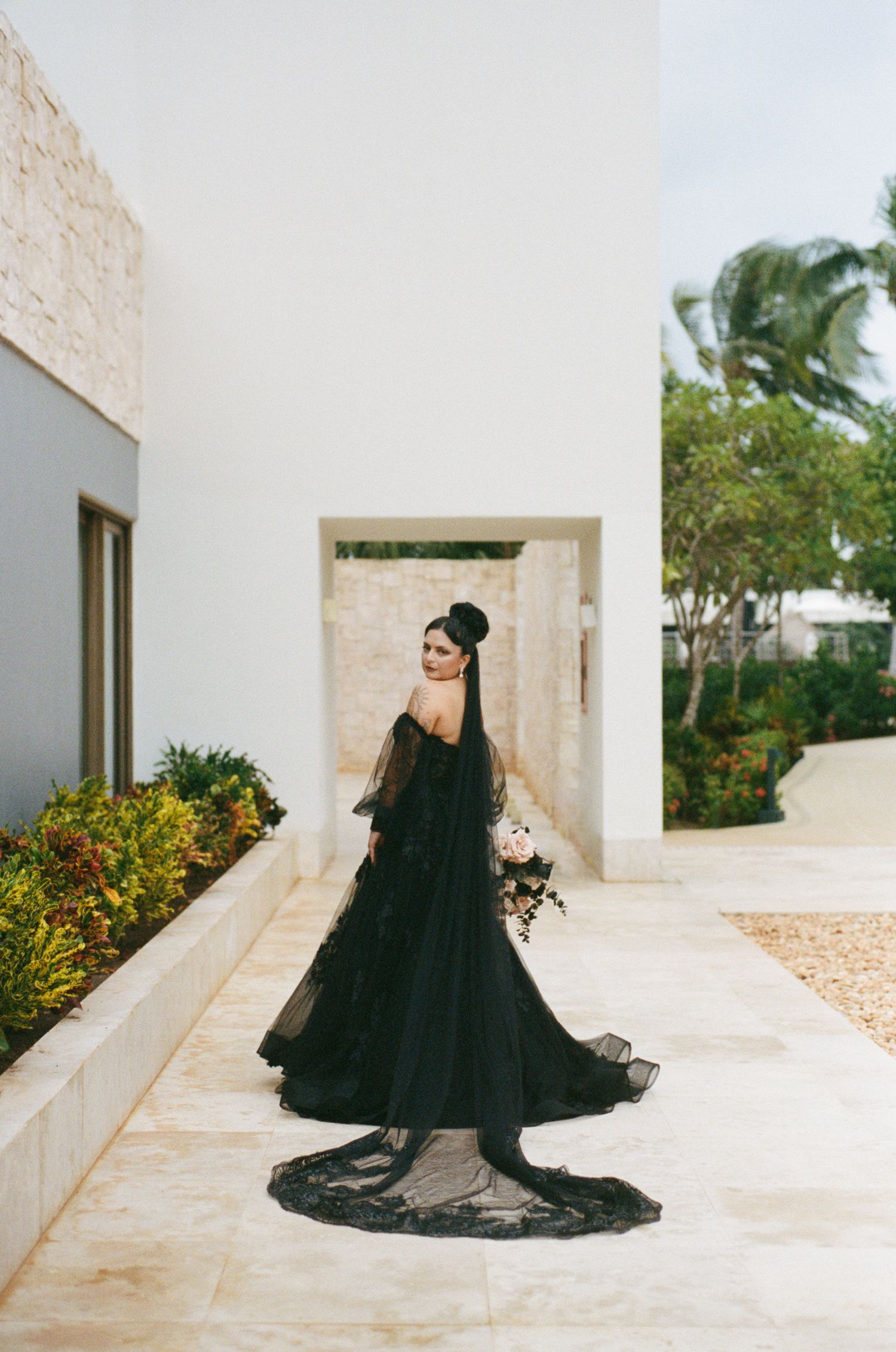 Bride in stunning black off-shoulder wedding gown with long lace train poses outdoors at modern venue.