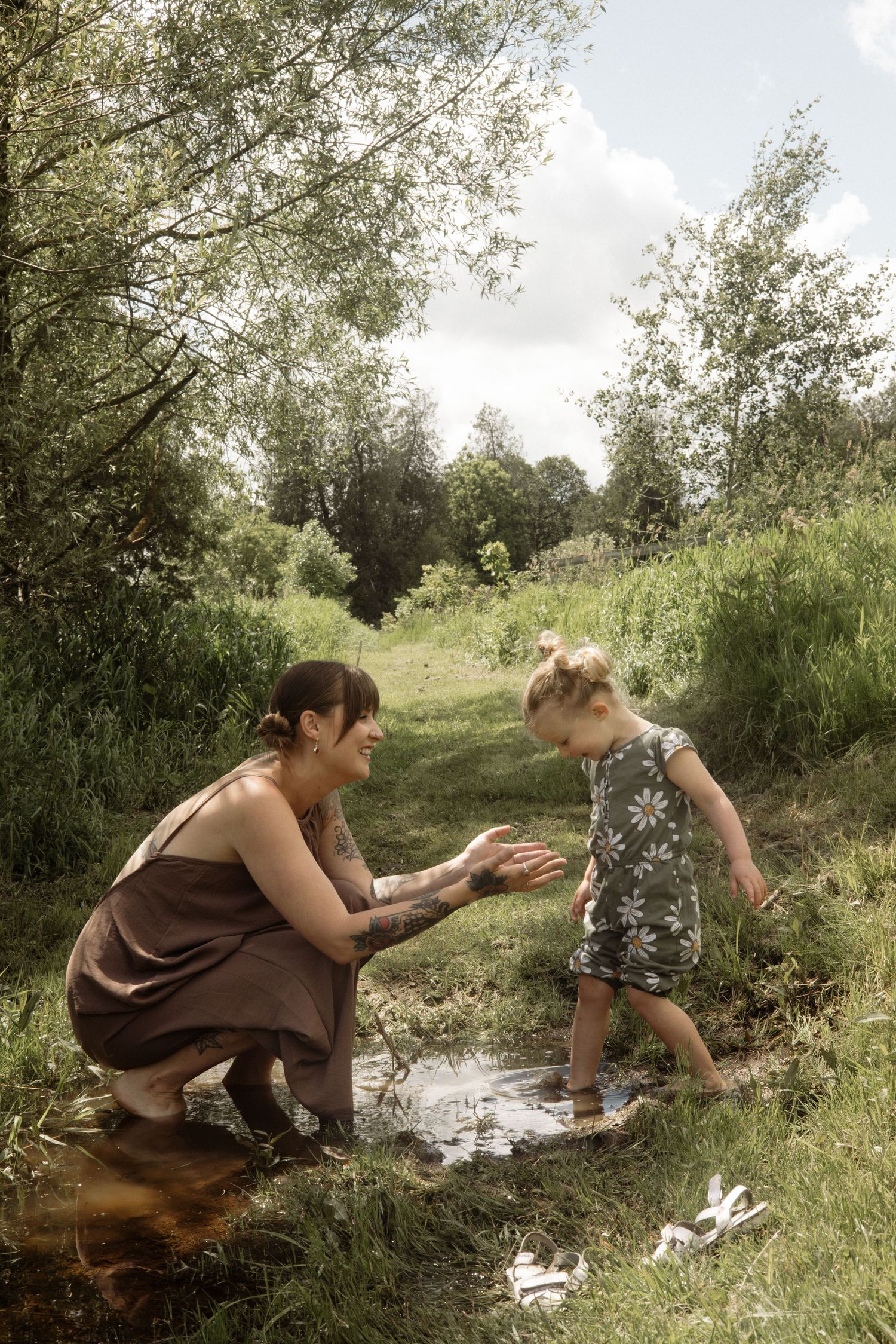 Tattooed mother crouching by a puddle holding hands with her smiling toddler in a daisy romper in nature.