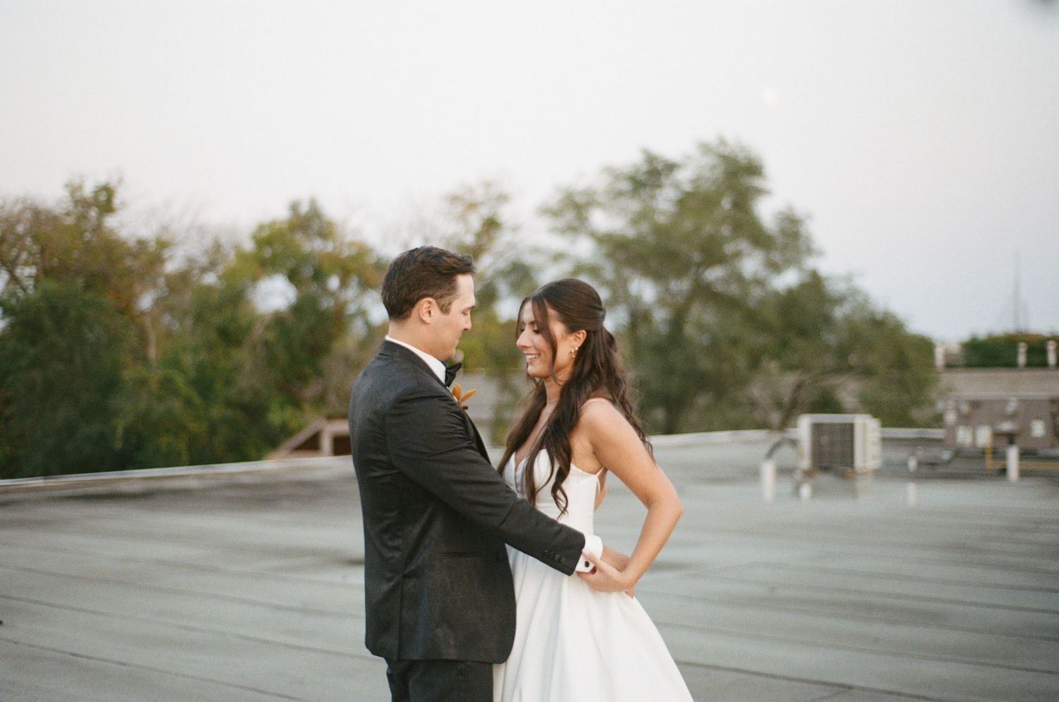 Smiling bride in white gown and groom in black tuxedo share a joyful moment on rooftop.