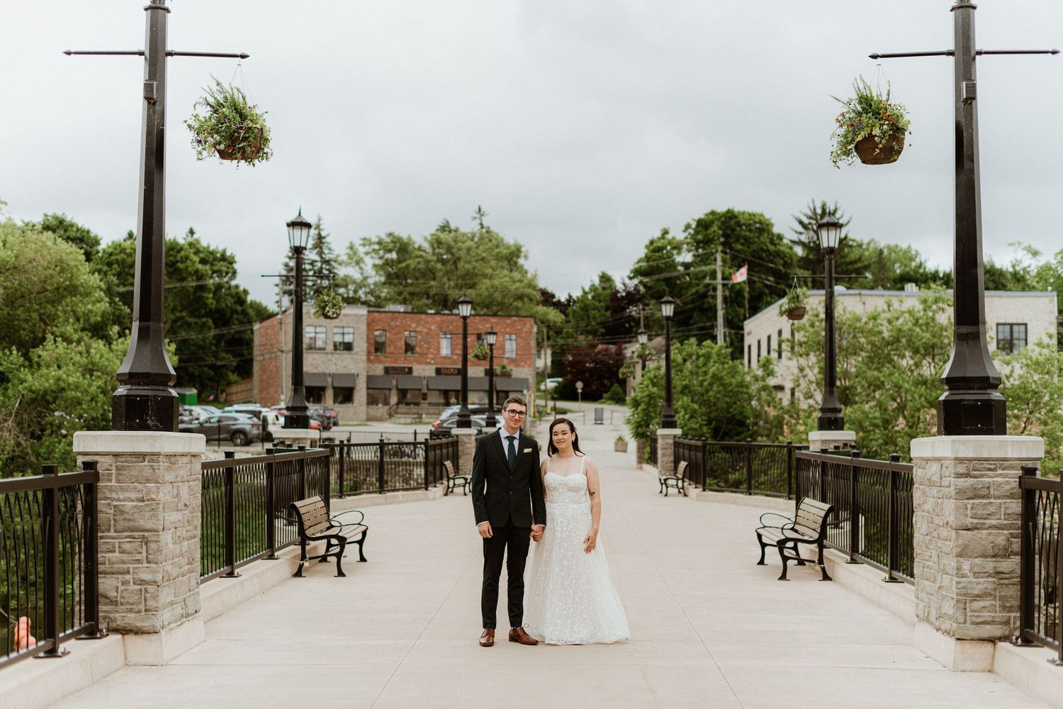 Wedding couple standing on bridge in small town with hanging flower baskets.
