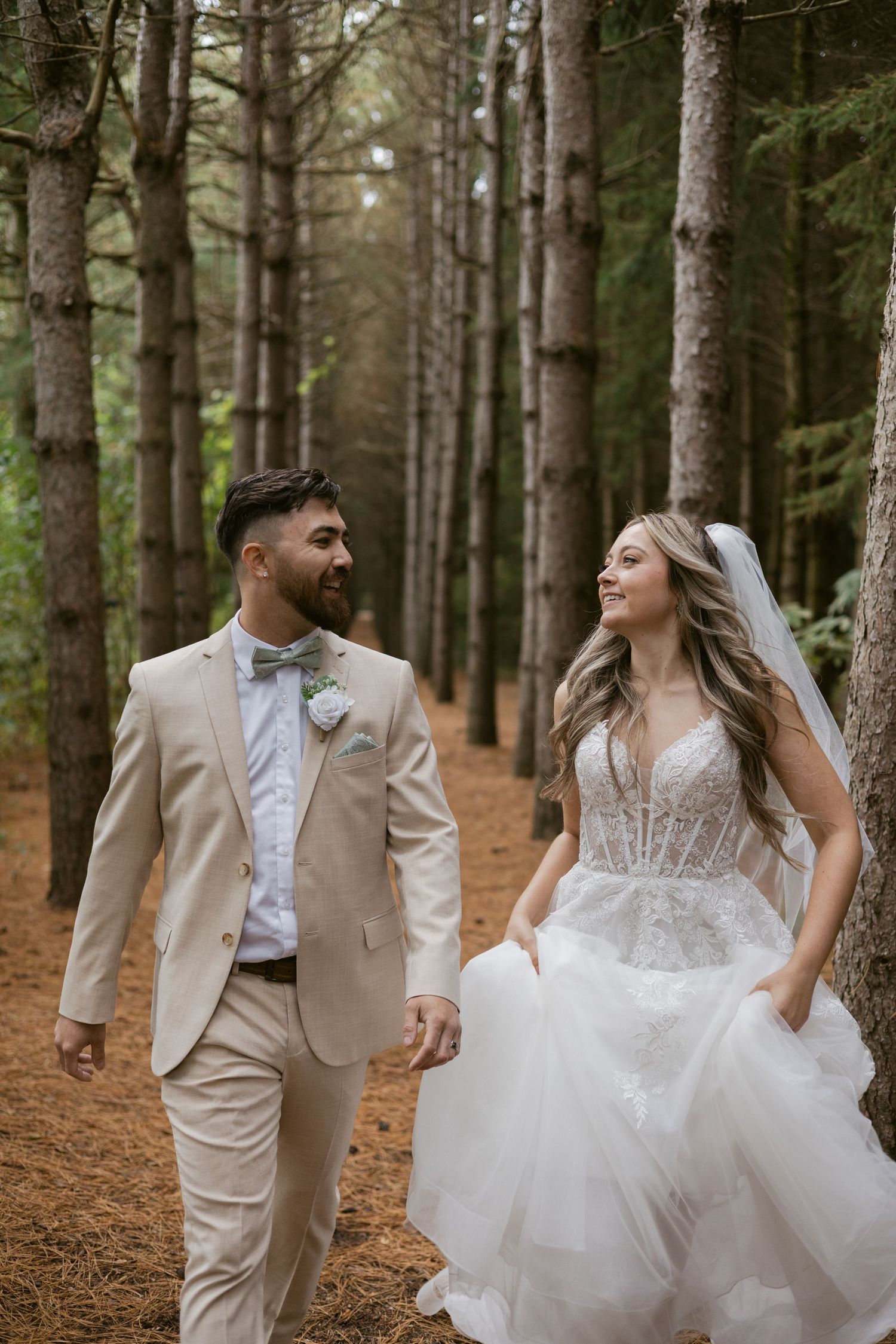 Happy bride and groom share a glance while walking through a tall pine forest wedding portrait.