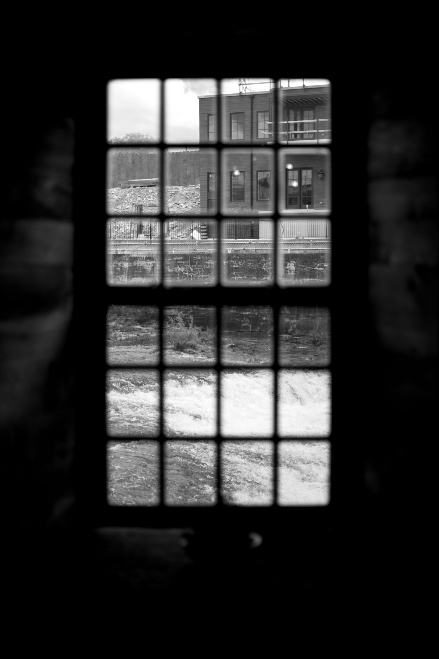 Black and white view through a multi-pane window overlooking a river and historic brick building.