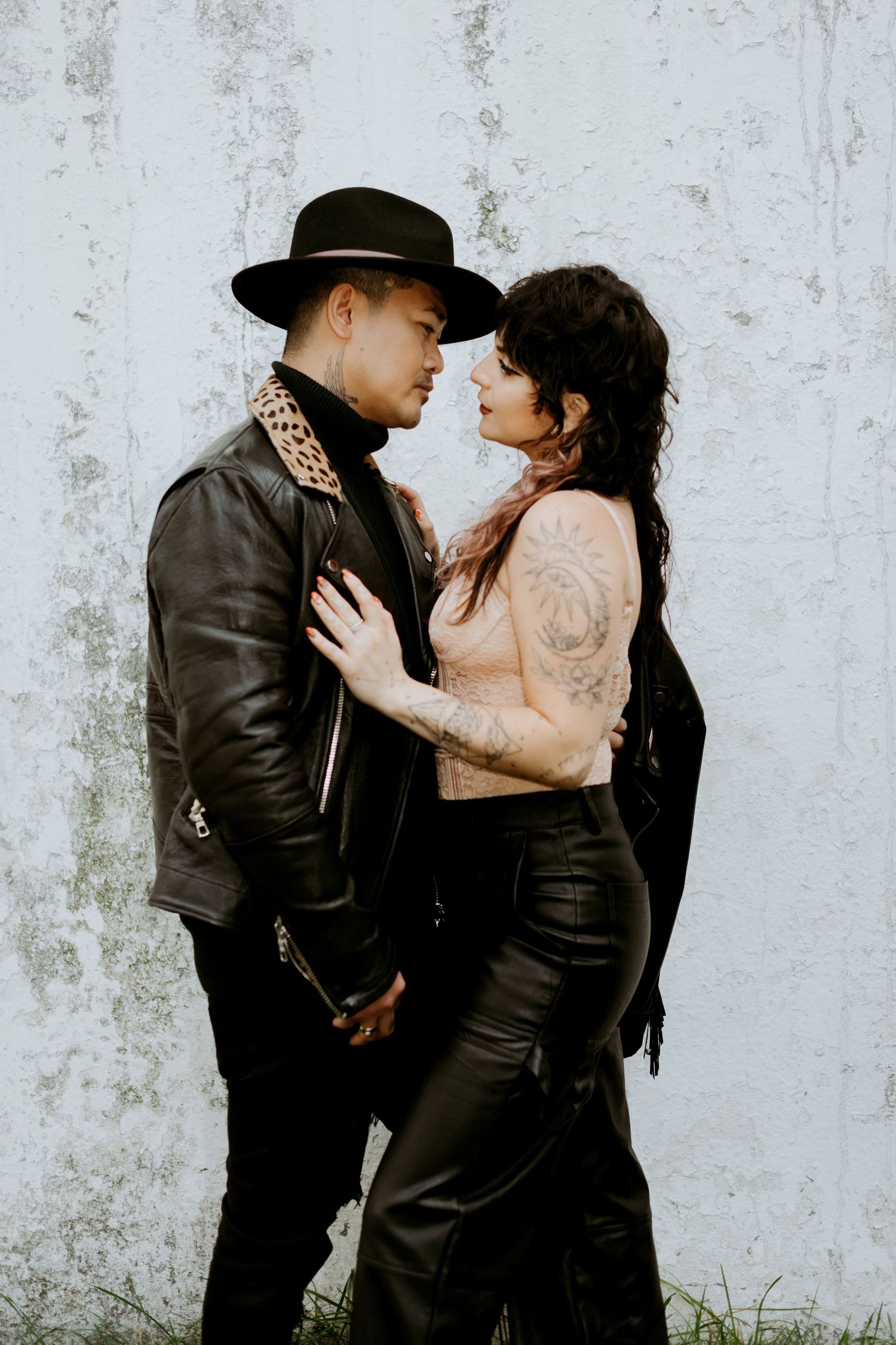 Tattooed couple in leather jackets sharing romantic moment against weathered wall.