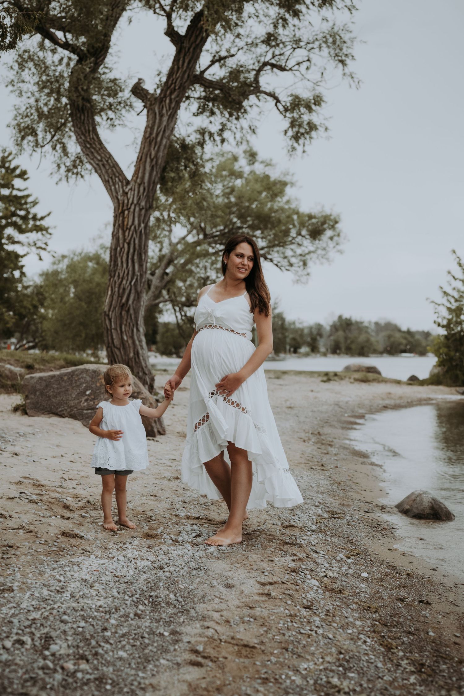 Smiling pregnant woman in white dress walks with toddler along a scenic shoreline.
