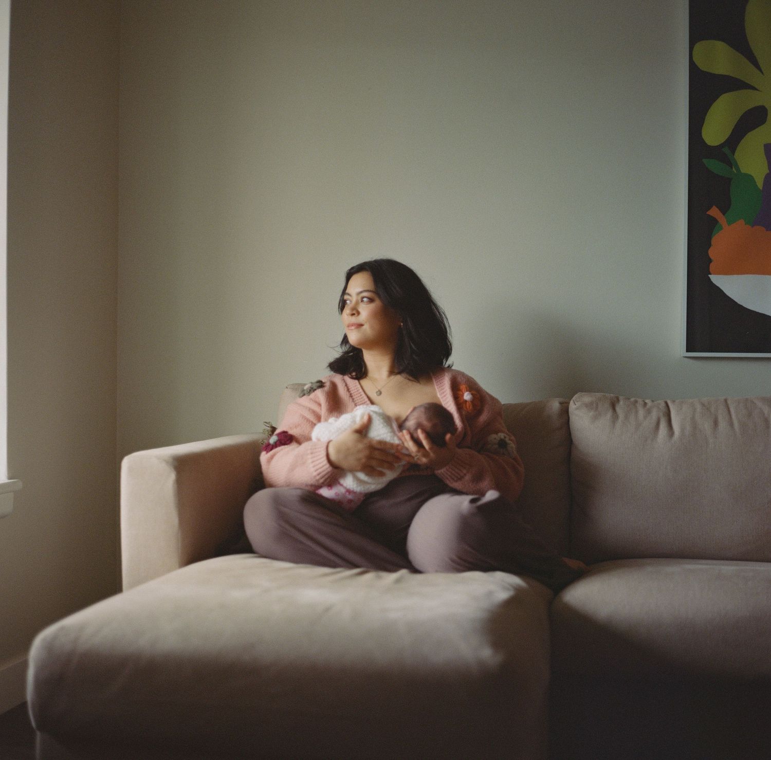 New mother sitting cross-legged on couch holding newborn baby while gazing out window.