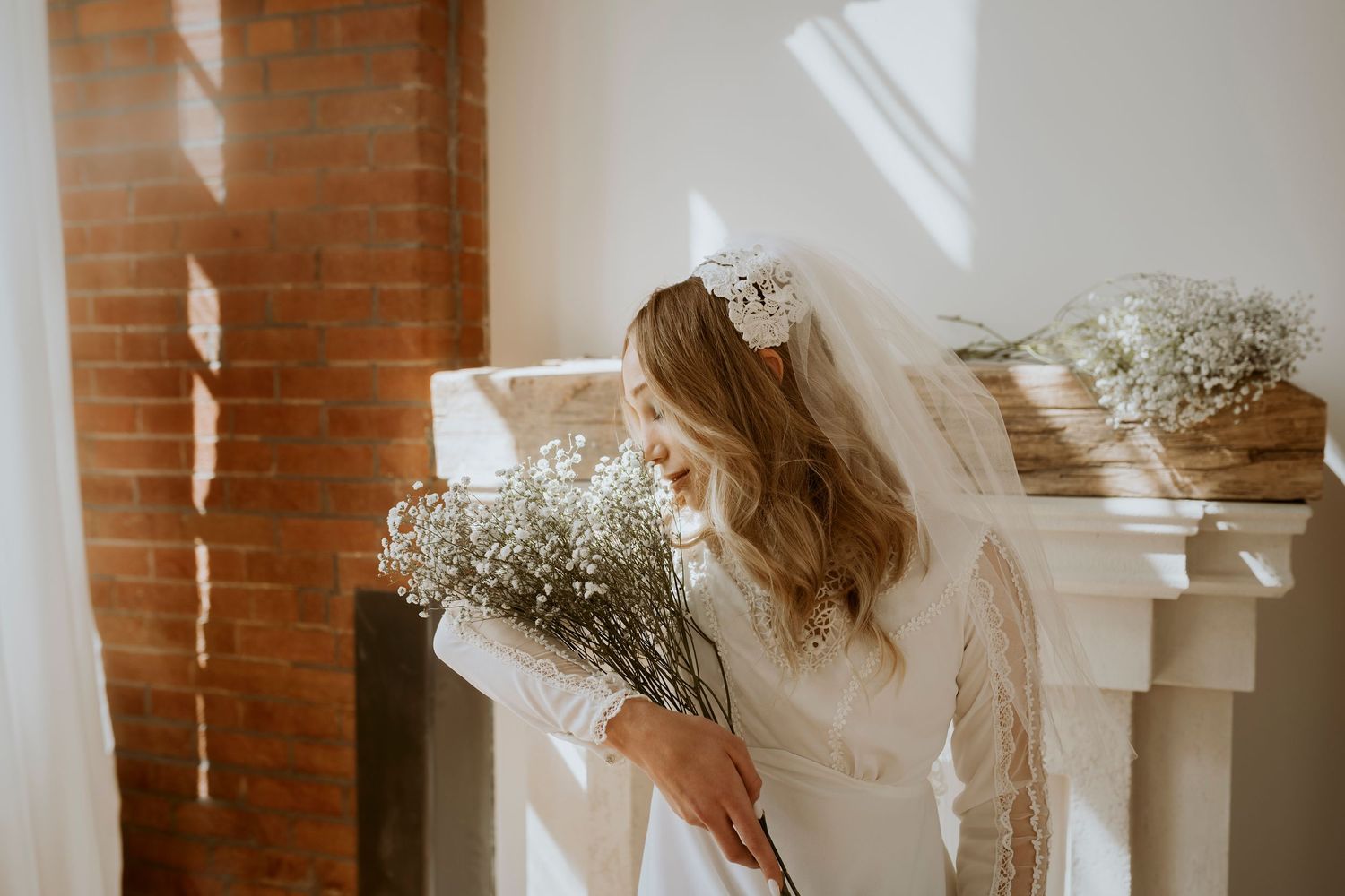 Bride in white lace wedding dress smiling while holding large baby's breath bouquet in sunlit rustic room with brick wall.