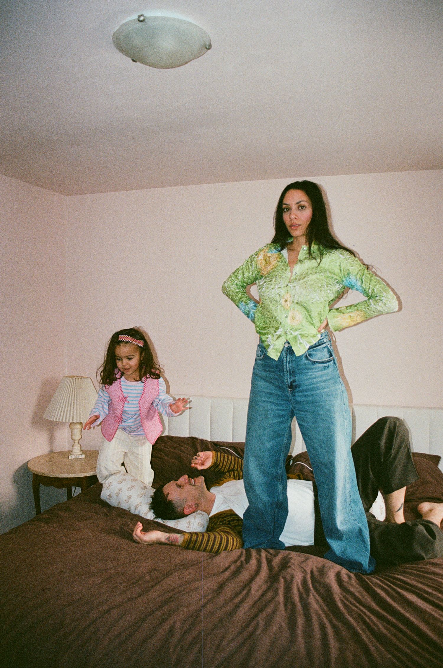Woman in green floral top stands on bed over man and young girl in pink vest in bright room.