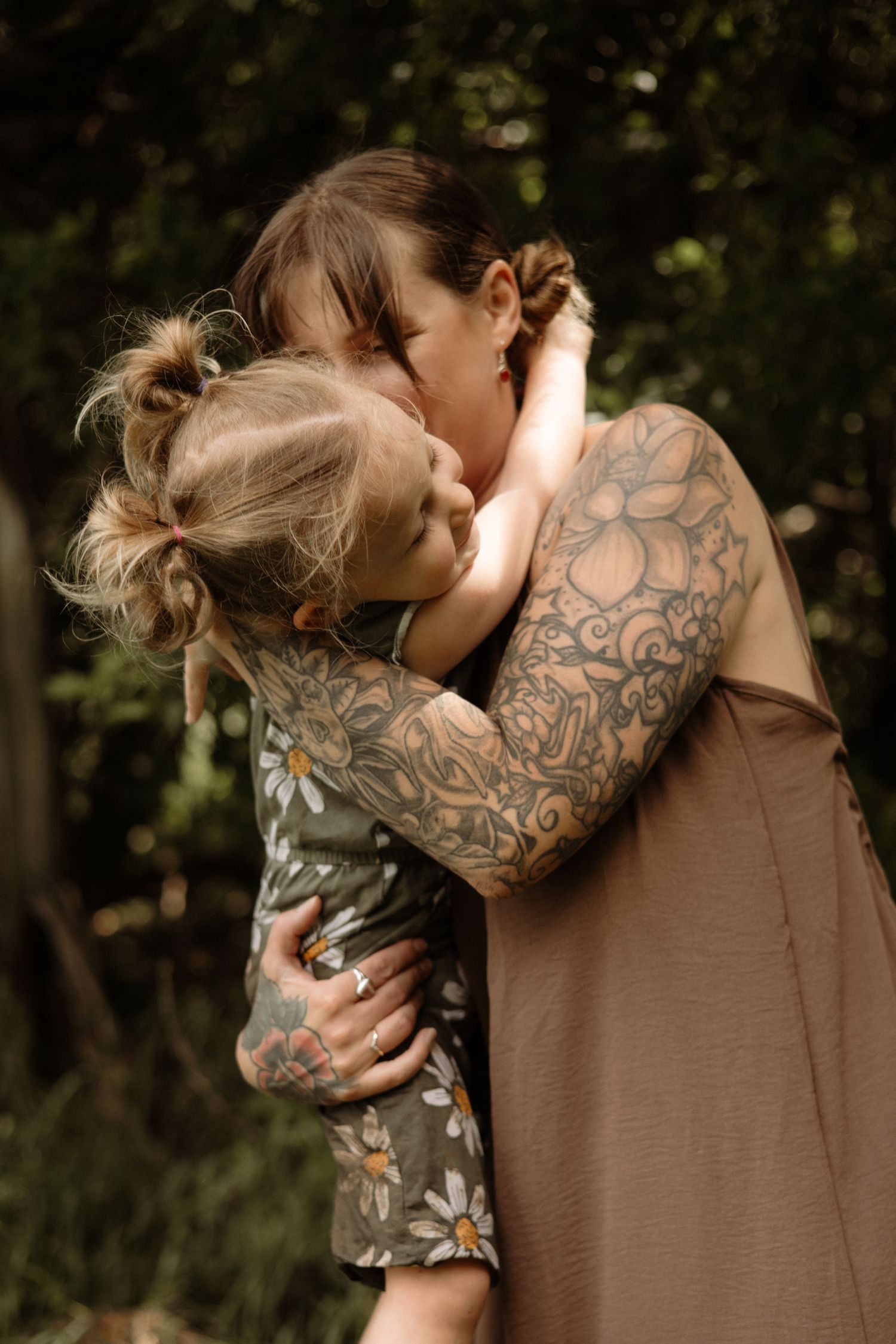 Tattooed mom hugs her toddler daughter wearing a daisy print dress outdoors.