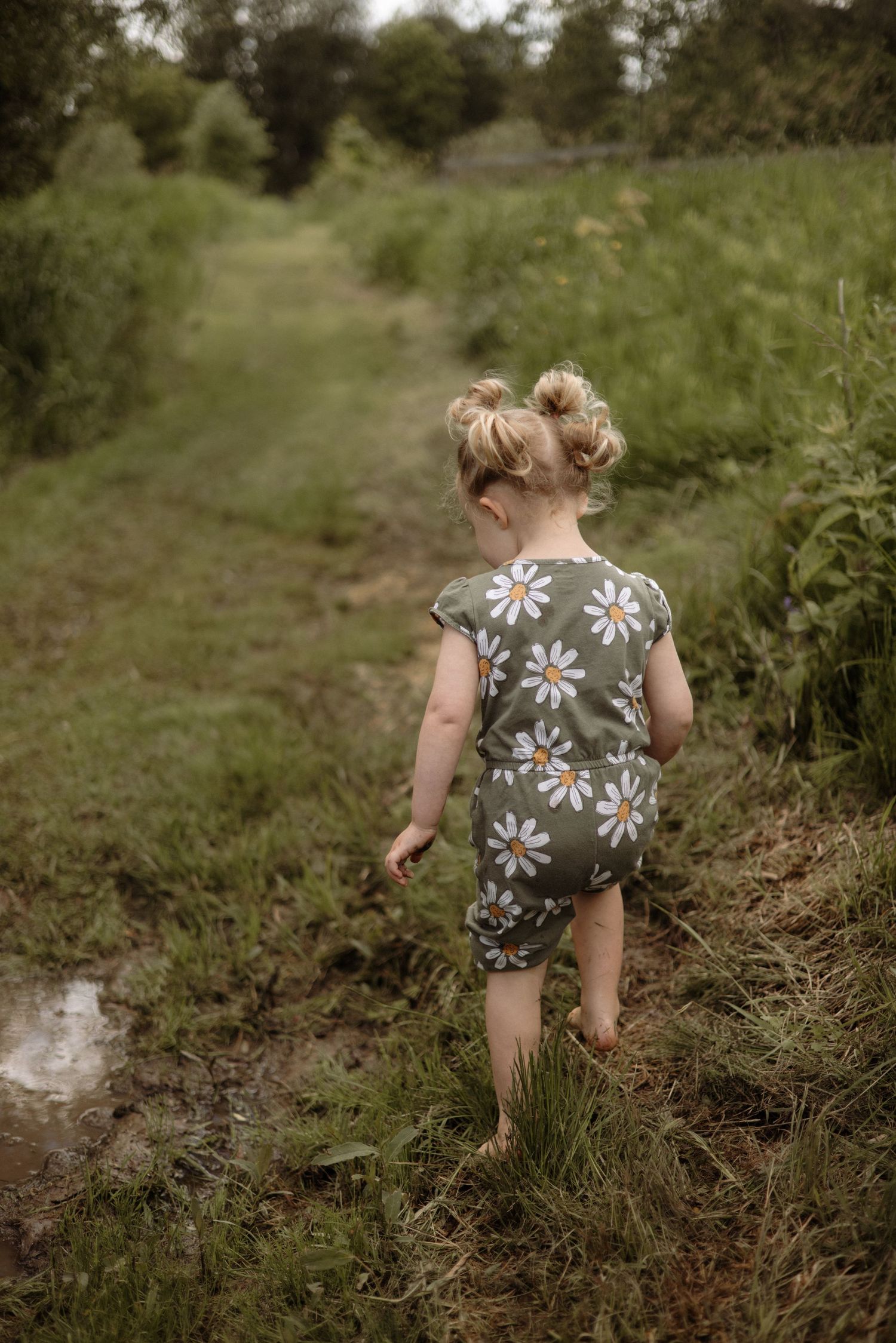 Barefoot toddler girl in daisy print romper walks along muddy path with green grass and puddles.