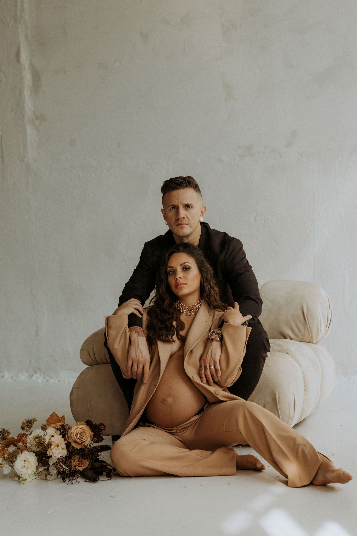 Couple maternity photo, man in black suit sits behind pregnant woman in beige outfit on white chair with flowers.