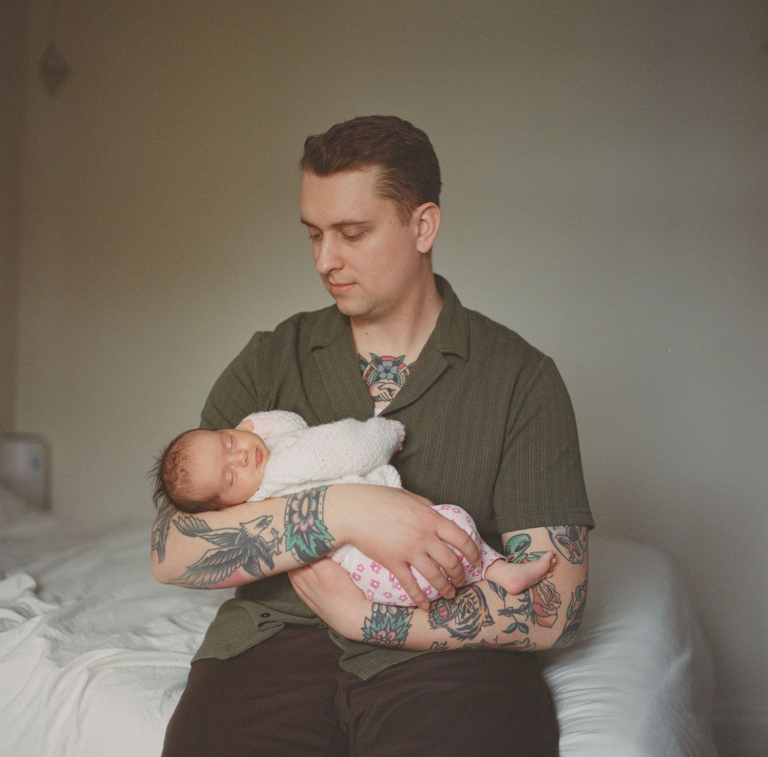 A tattooed father in a green shirt gently cradles his sleeping newborn baby while seated on a bed.