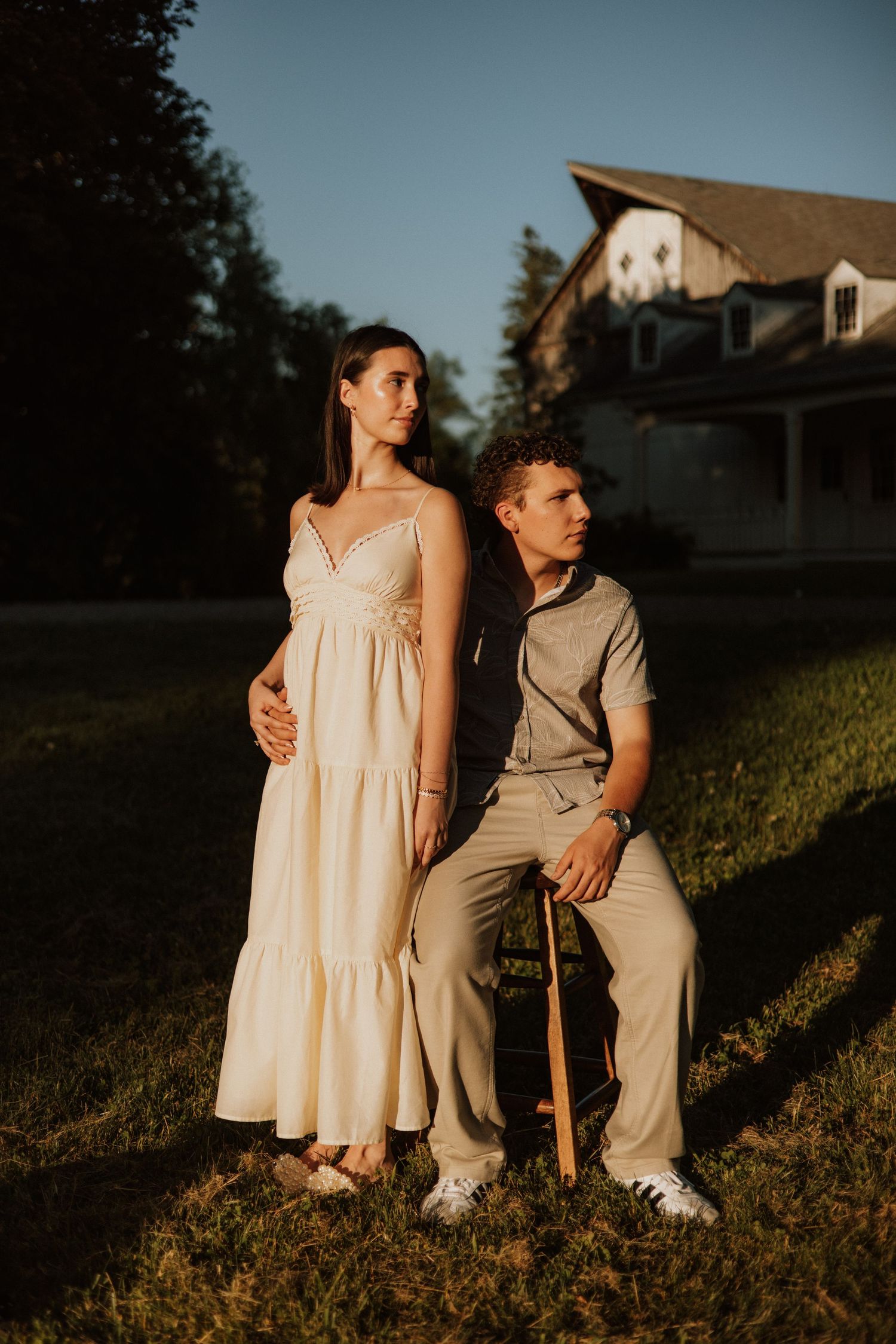 Pregnant couple poses outdoors at golden hour, woman in white maxi dress, man seated on chair, with a house in the background.