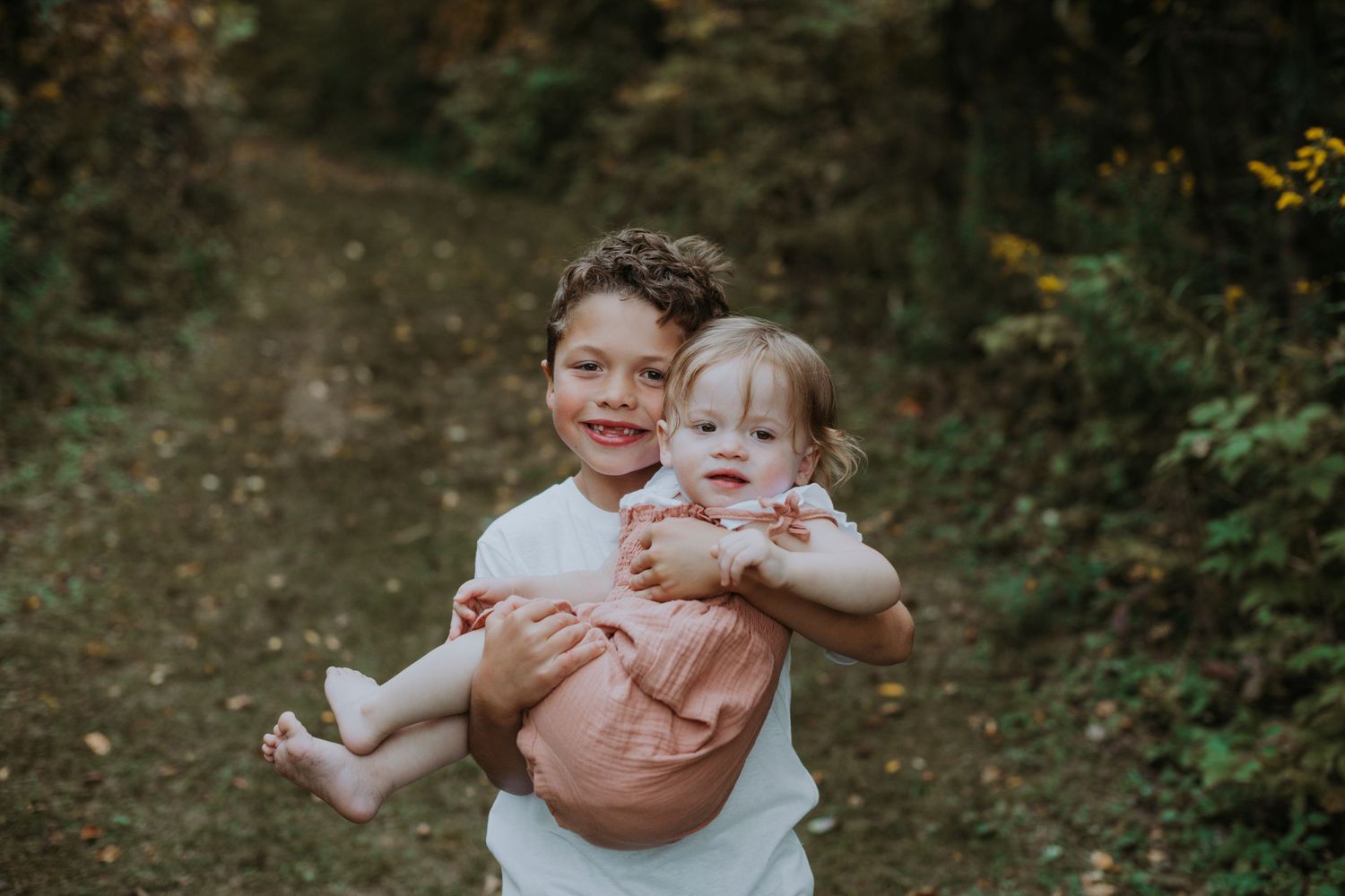 Smiling boy hugging his baby sister in a pink romper on an autumn forest trail.