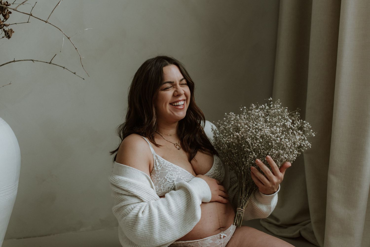 Smiling pregnant woman in white lace lingerie and cardigan holds baby's breath flowers in a studio.