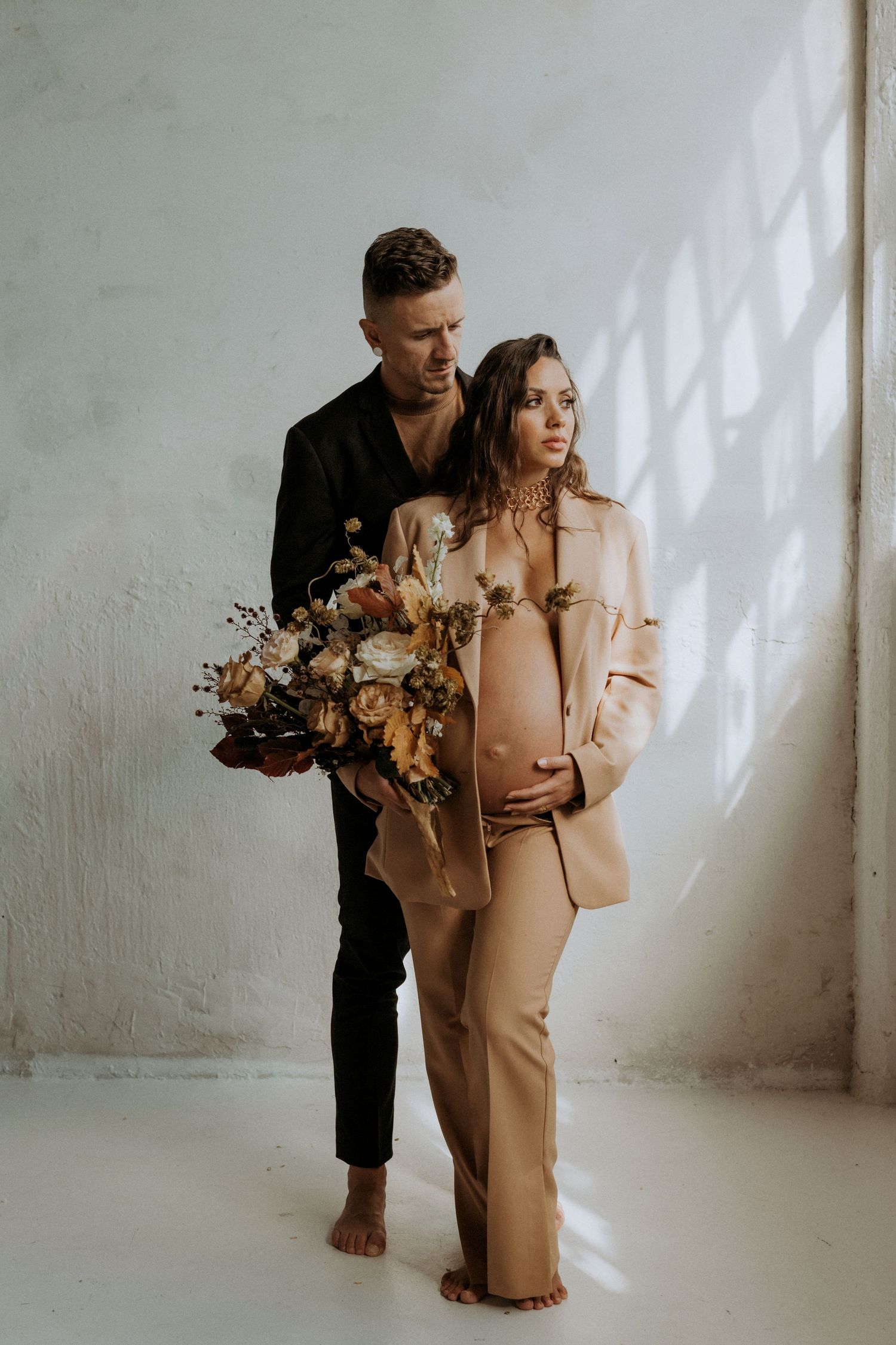Couple maternity portrait, man in black holds floral bouquet embracing pregnant woman in beige blazer in studio.