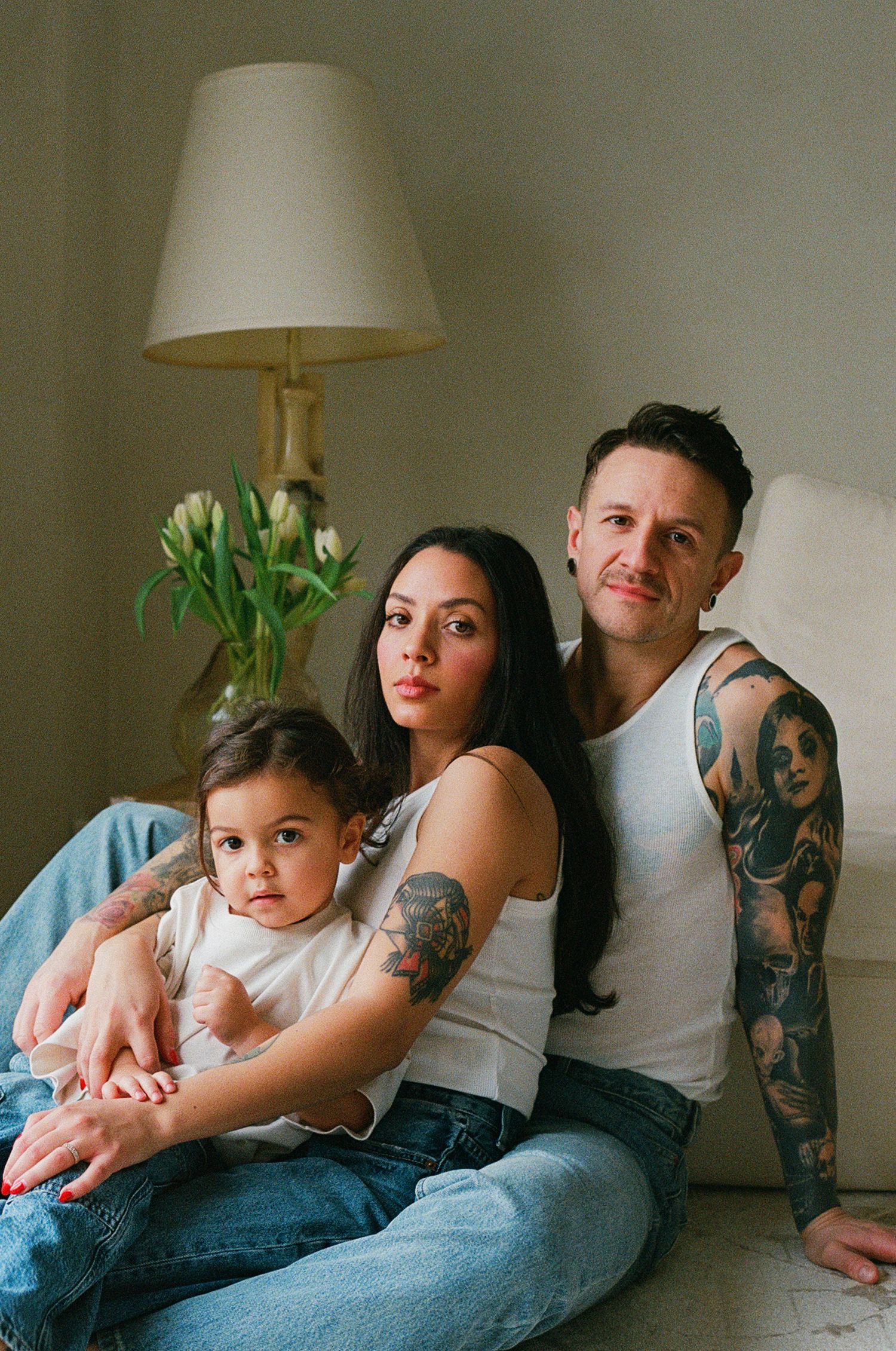 Tattooed family portrait with parents and toddler sitting on floor by lamp and tulips.