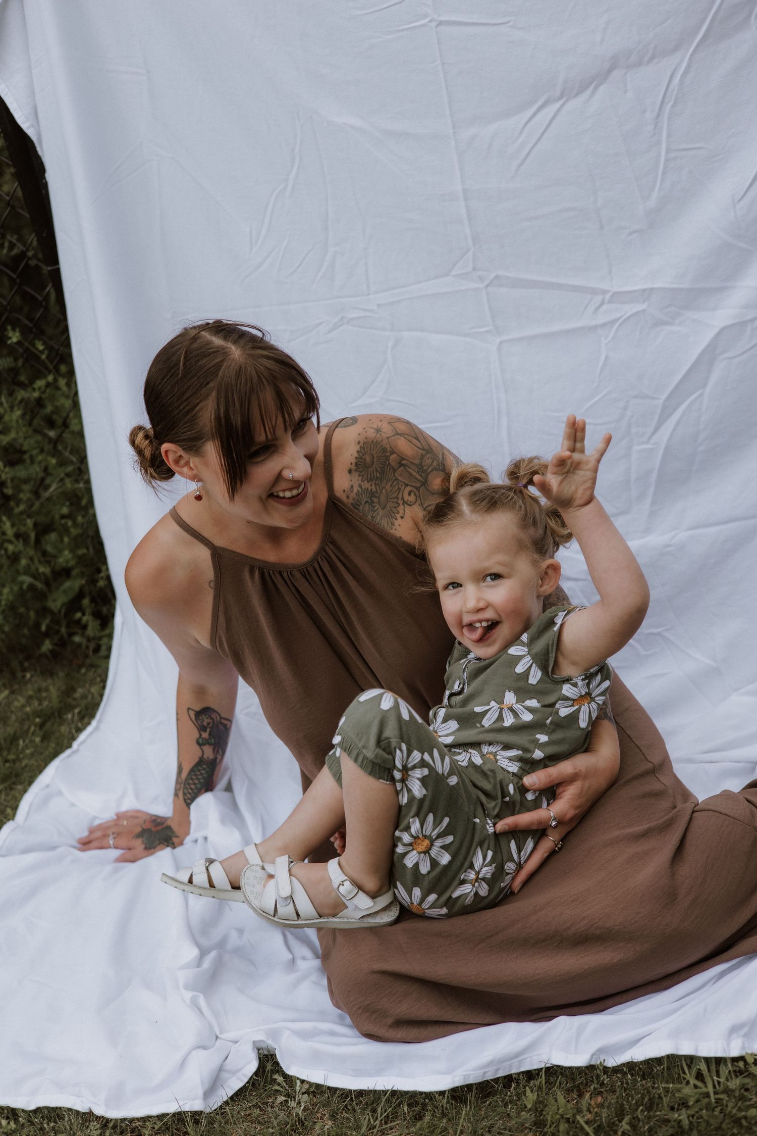 Laughing mother with tattoos sits on white blanket outdoors with excited toddler girl in floral romper.