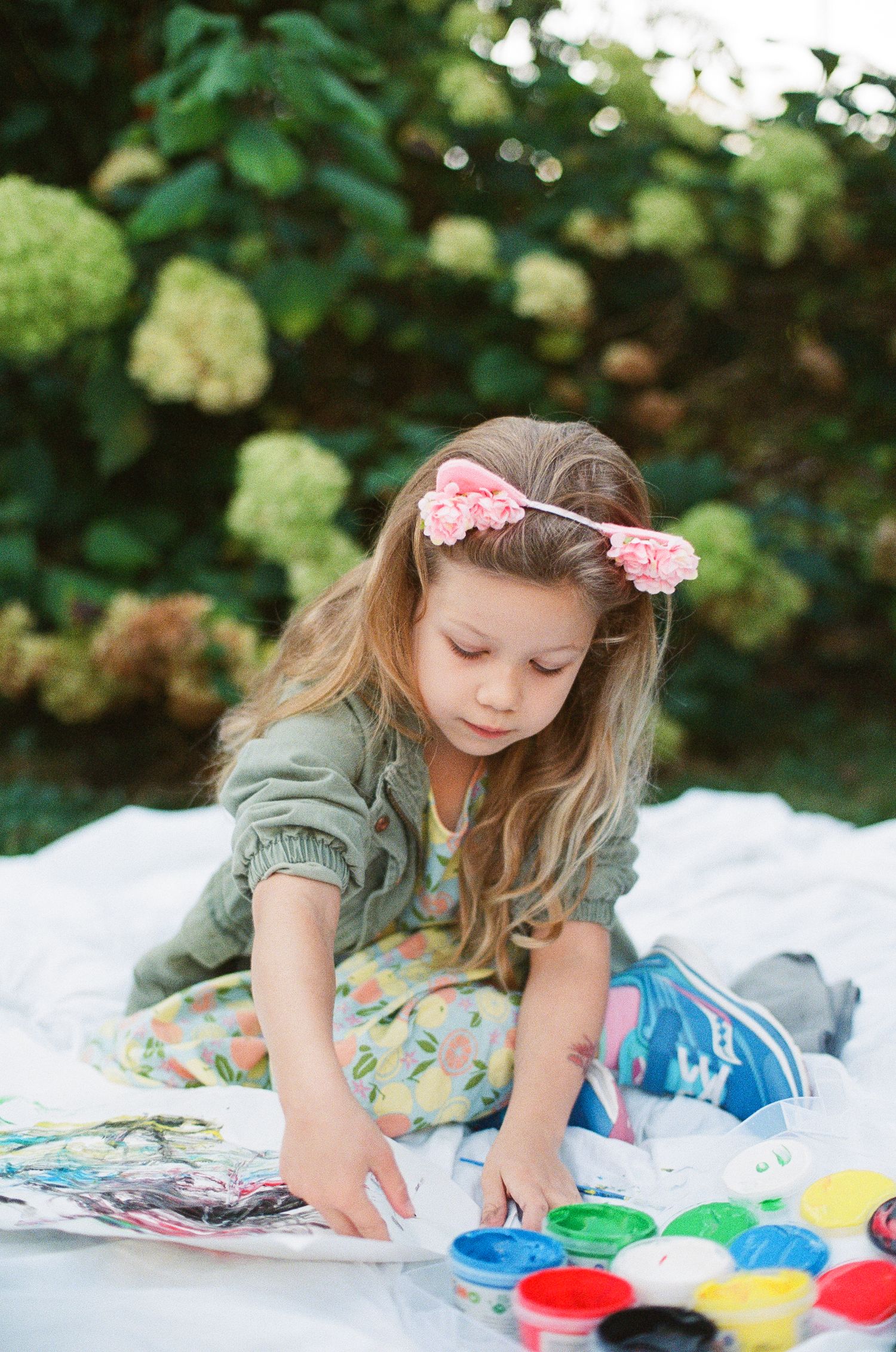Young girl with pink flower crown painting with colorful paints outdoors near hydrangeas.