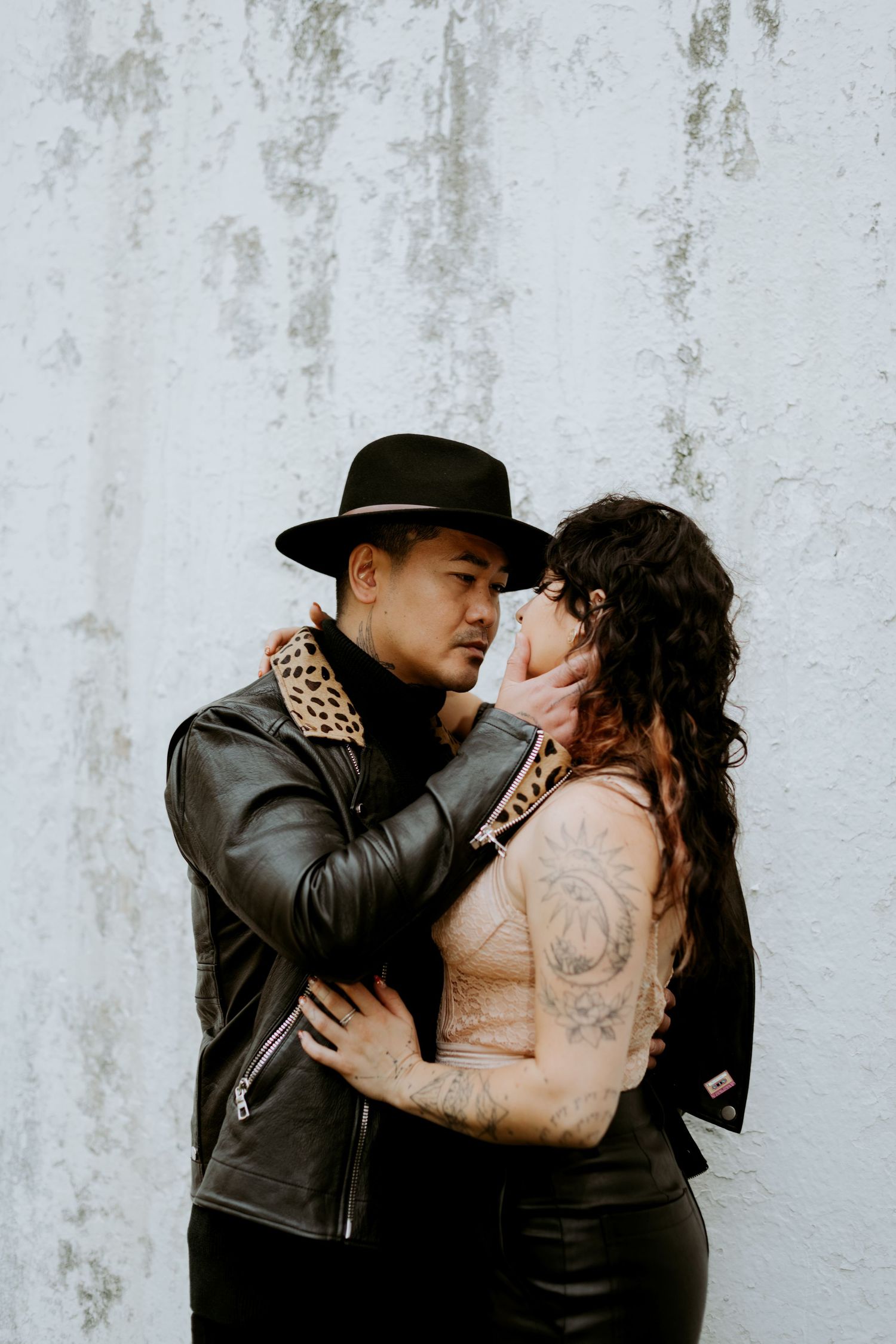 Couple in black leather jackets embracing intimately against a distressed white wall.