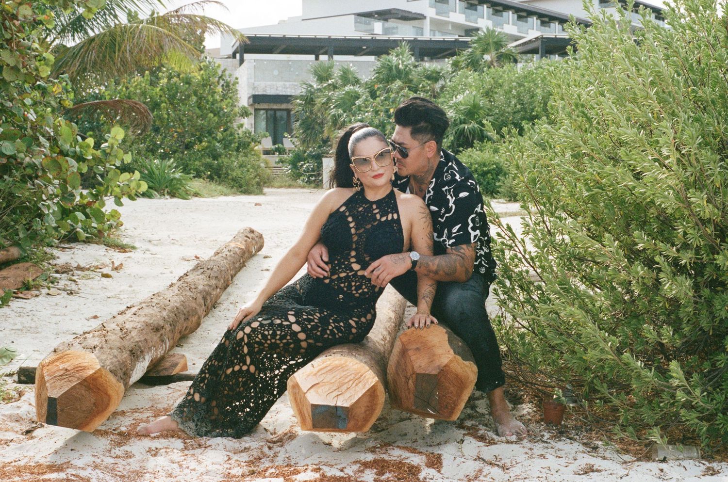 Couple posing on logs in a tropical garden, both wearing black outfits with sunglasses.