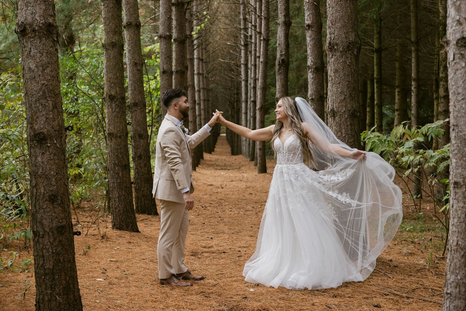Groom twirls bride in a white lace gown with flowing veil in a pine forest setting.