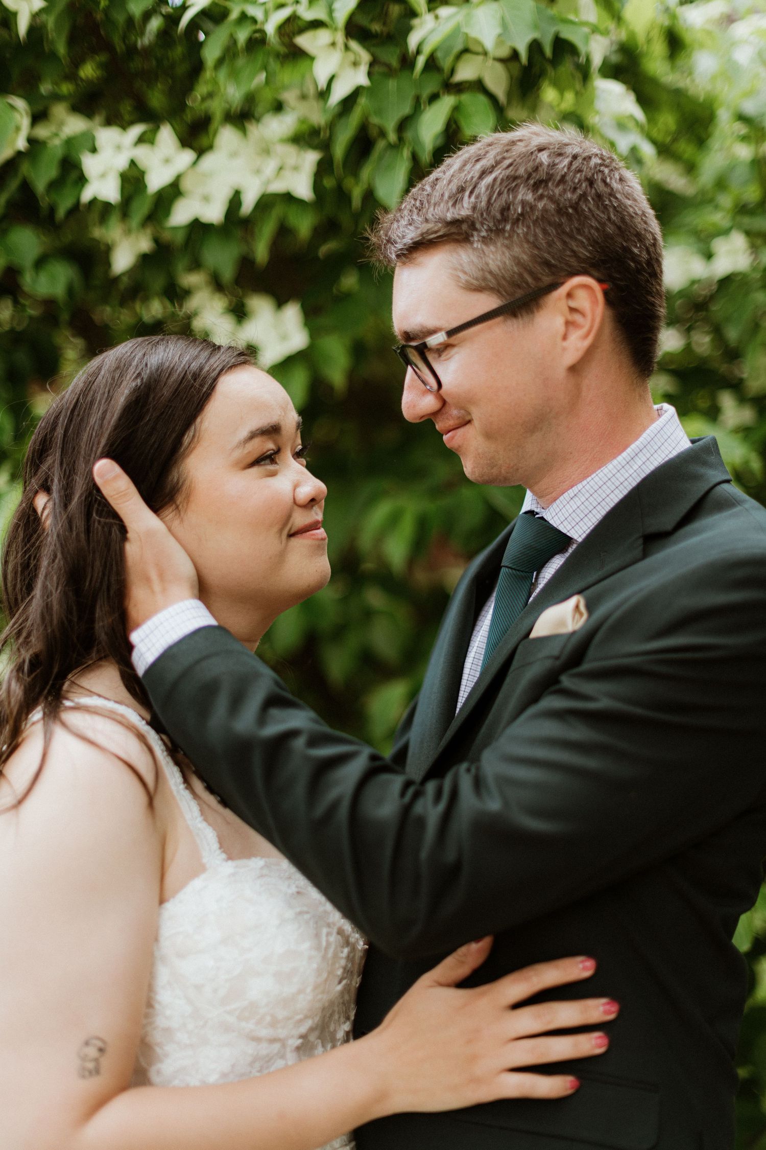 Groom tenderly cups bride's face as they share a close romantic moment among green leaves.