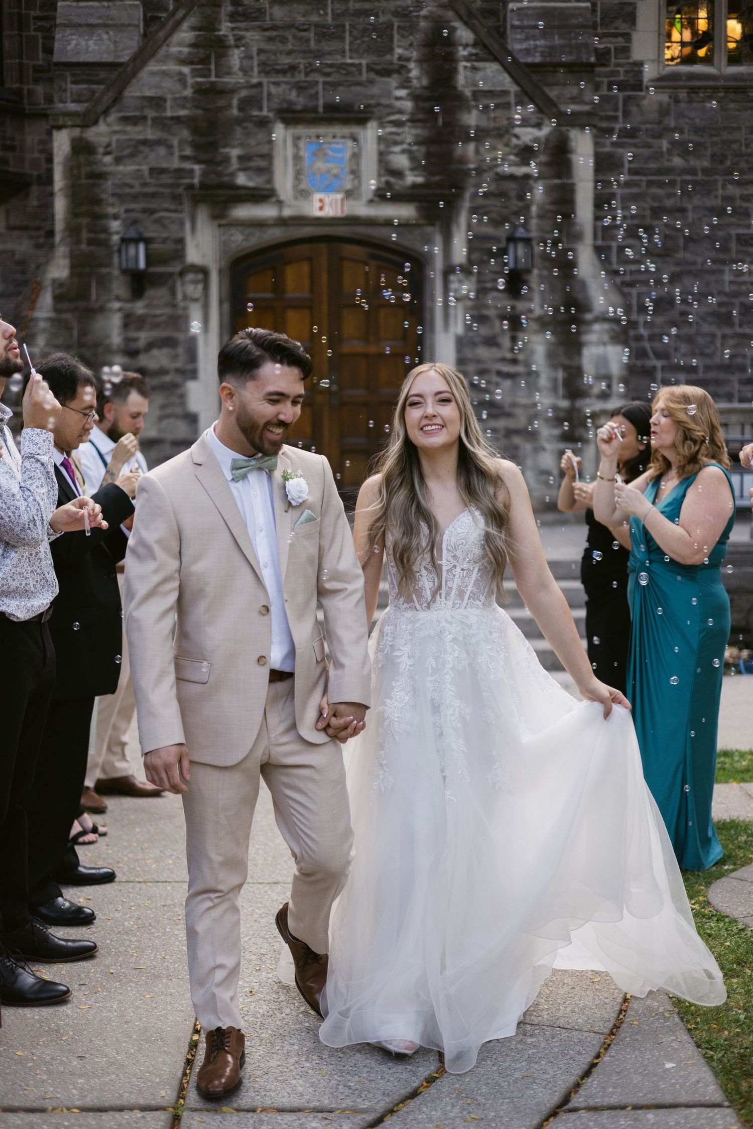 Newlyweds walk hand-in-hand through bubble send-off as guests cheer outside gothic stone church.