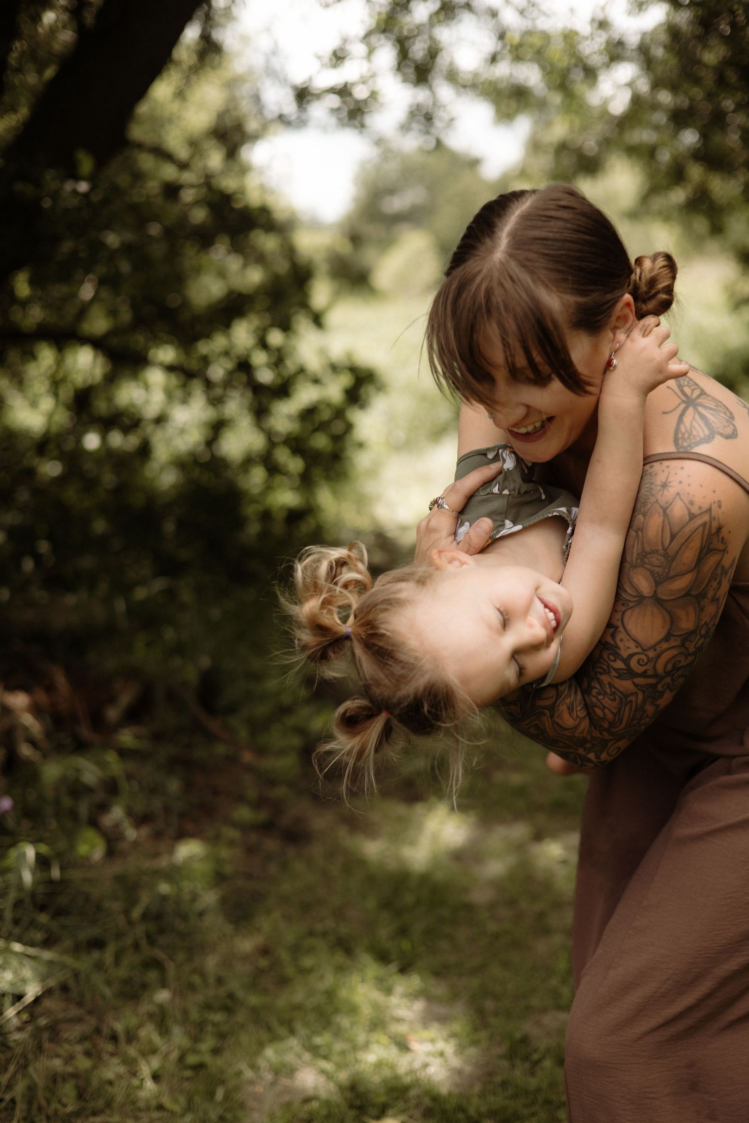 A tattooed mother playfully swings her giggling toddler upside down outdoors.