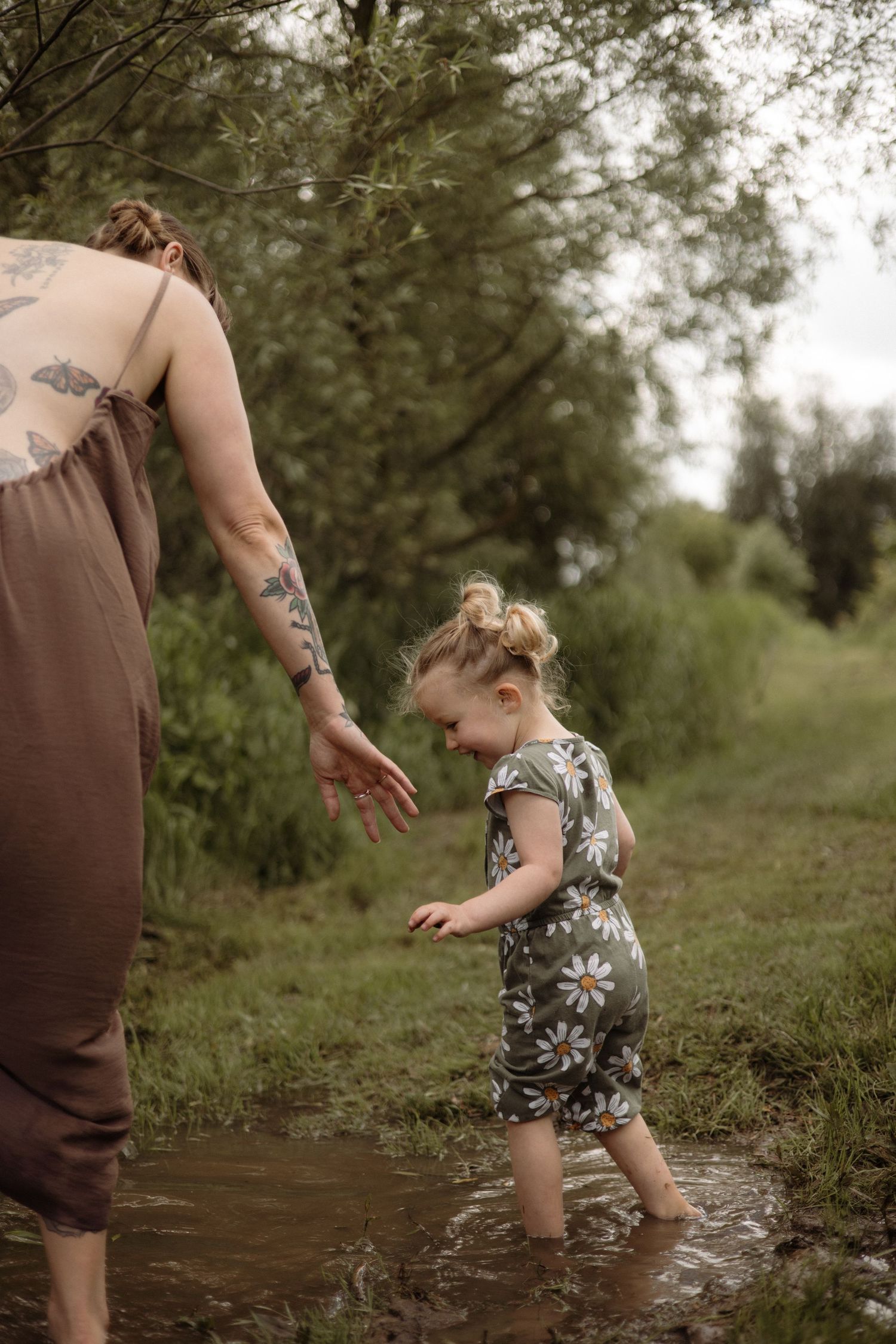 Tattooed mom reaches out to her smiling toddler in a floral romper splashing in a muddy puddle.