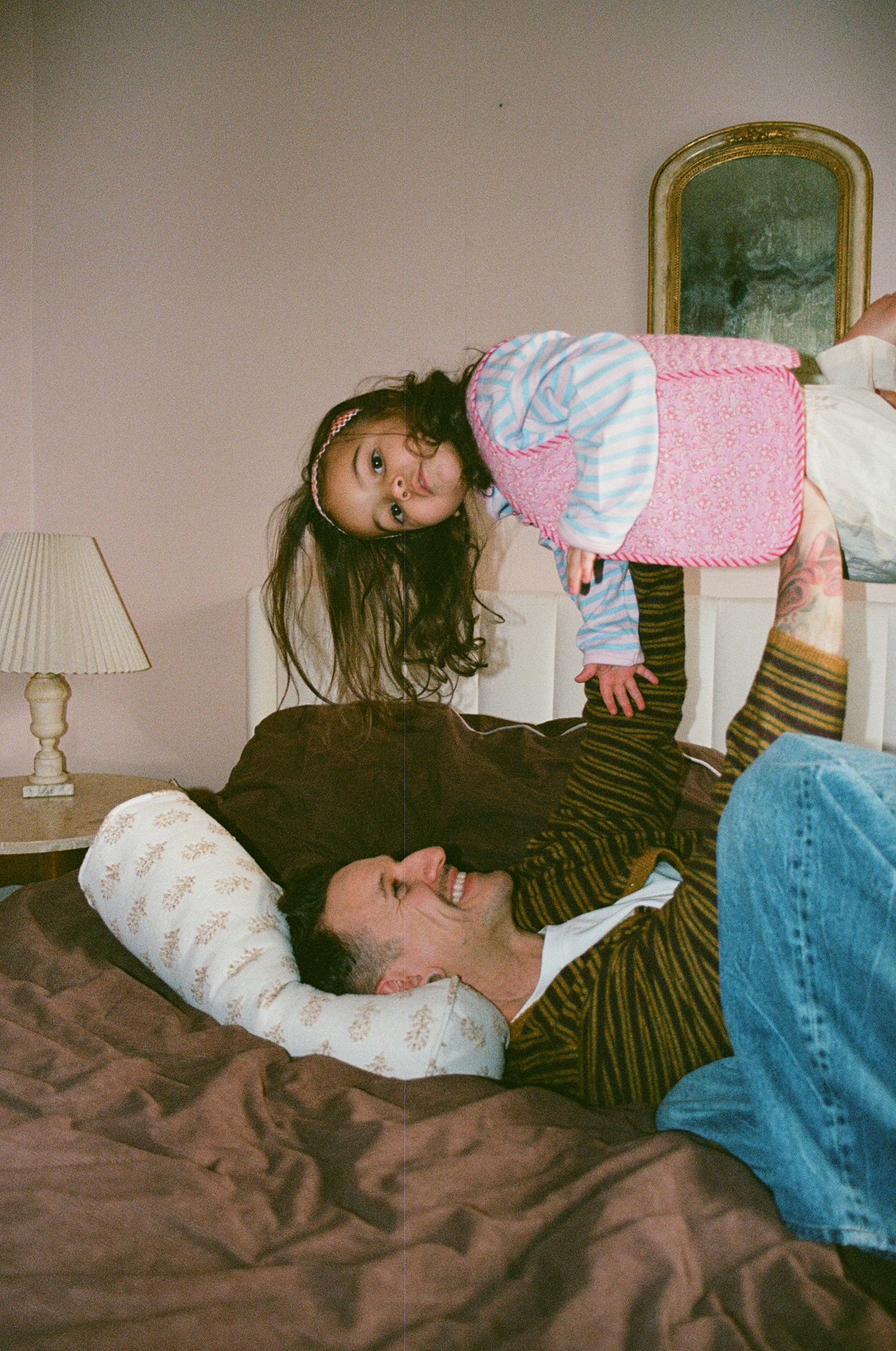 A young girl stands on a man lying on a brown bed in a cozy bedroom with a lamp and mirror.