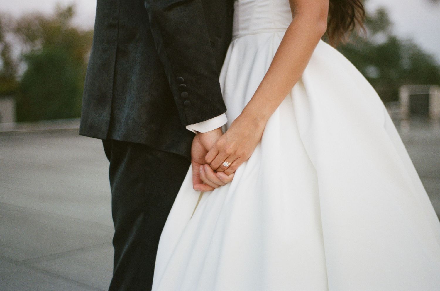 Close-up of bride and groom holding hands, bride wearing a flowing white ball gown on a rooftop.