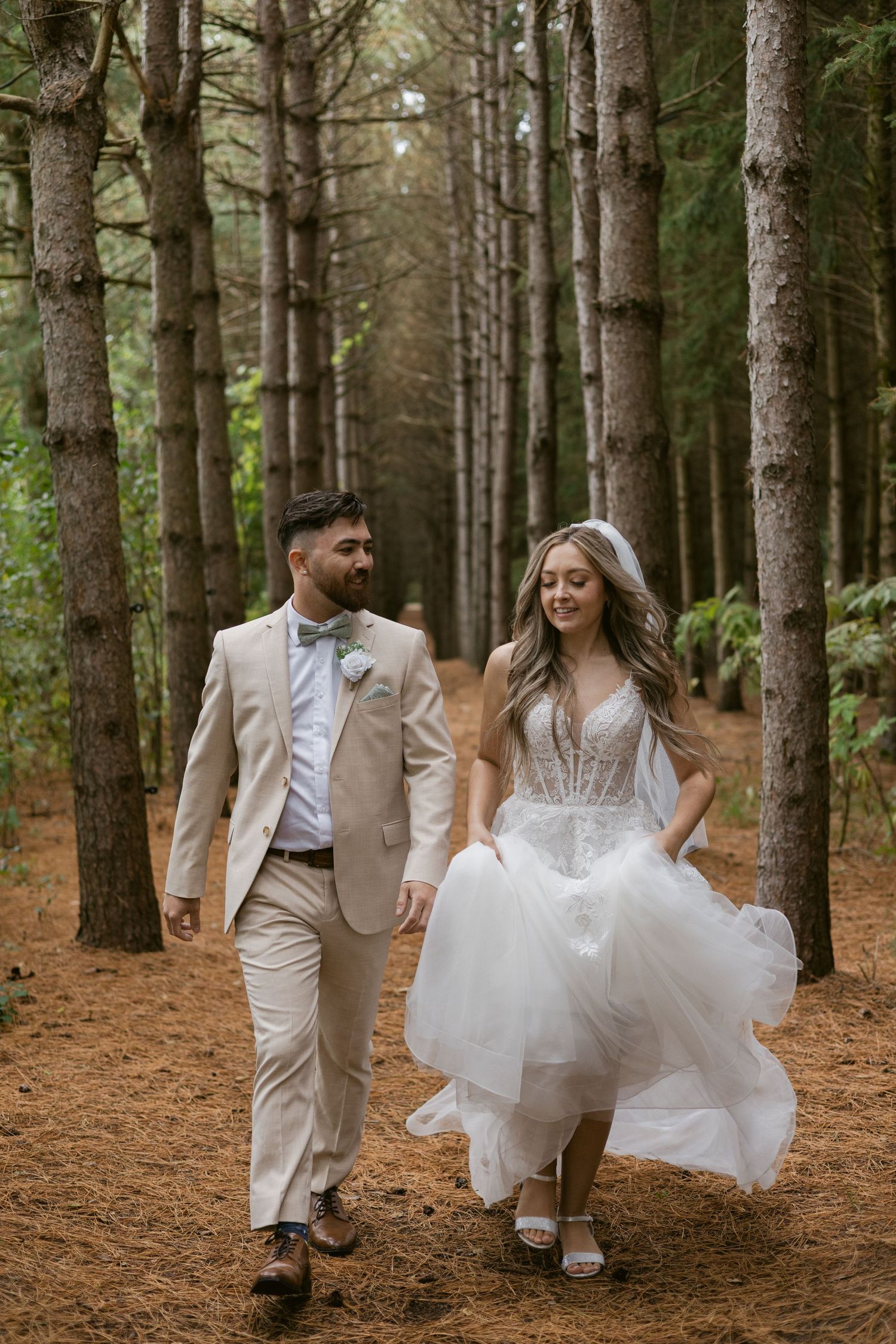 Bride and groom stroll through a pine forest path, groom in tan suit and bride in white gown.