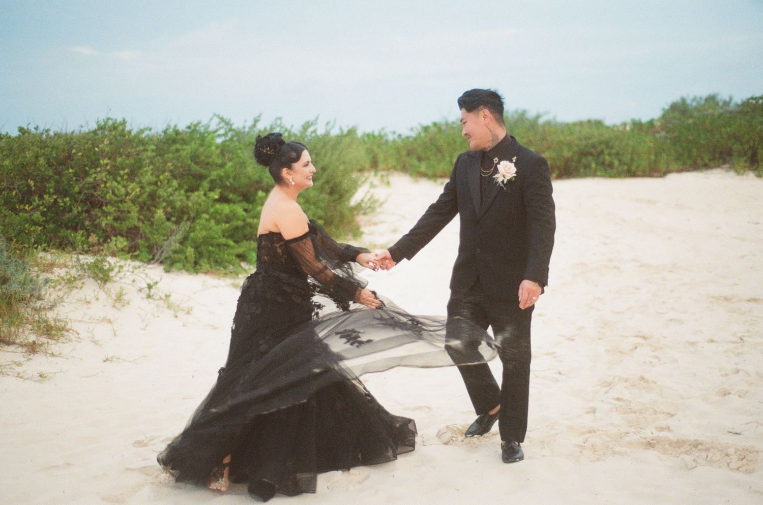 Wedding couple in all-black attire hold hands facing each other on white sand beach.