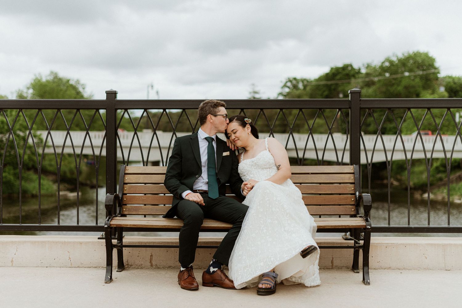 Romantic bride and groom share a tender moment on a park bench by a scenic river.
