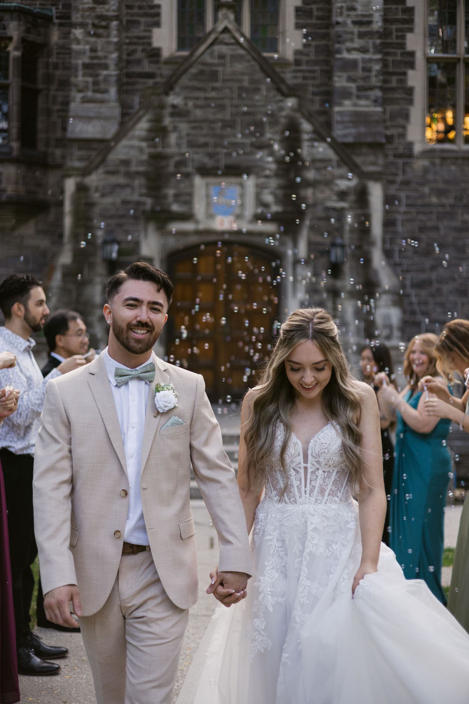 Smiling bride and groom walk hand-in-hand through bubble send-off outside historic stone chapel.