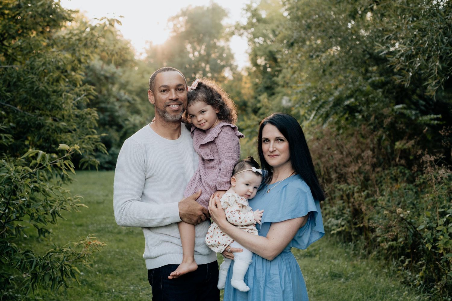 Happy family of four posing outdoors with two young children among lush green trees.