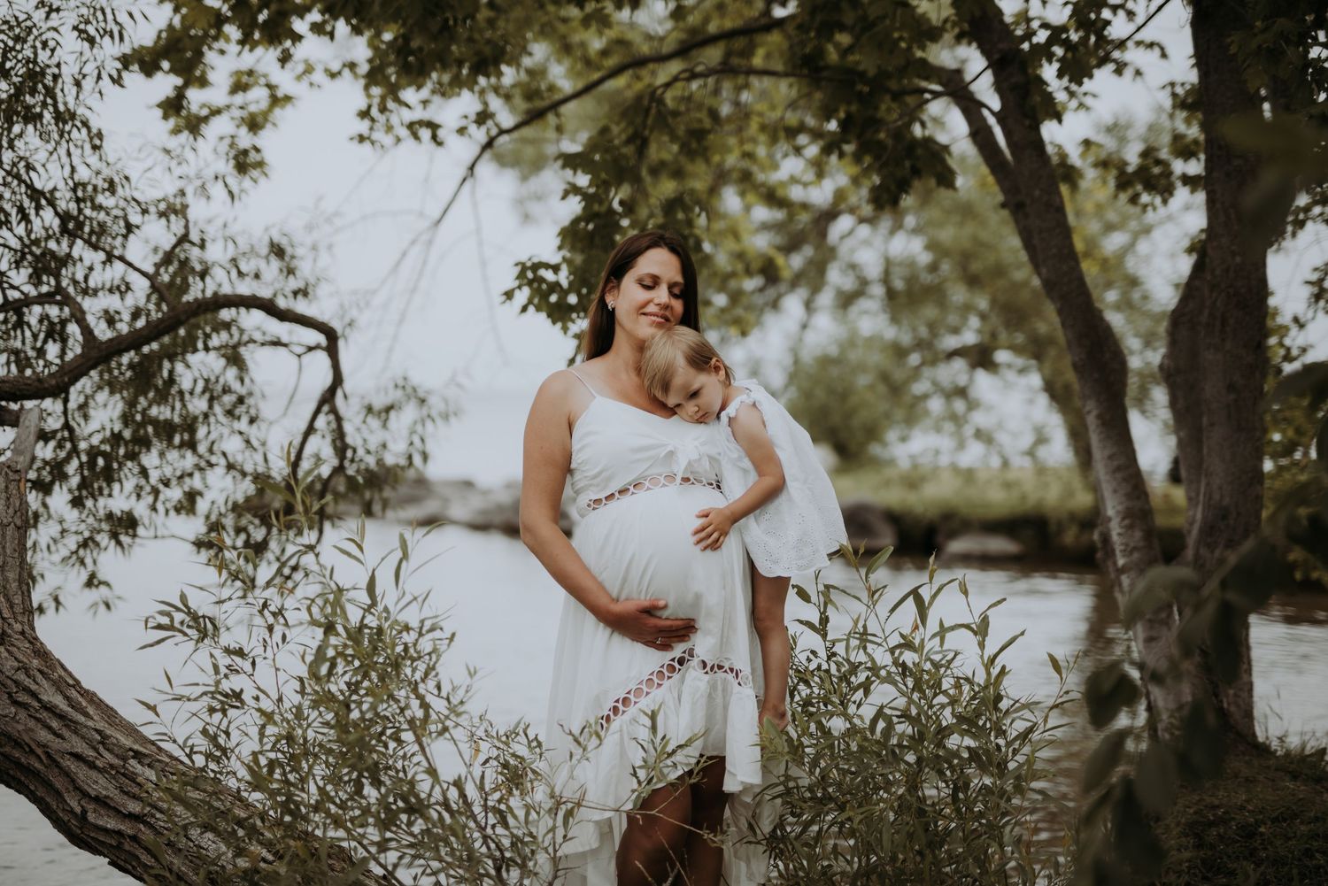 Pregnant woman in white dress embraces child near trees by a tranquil river.