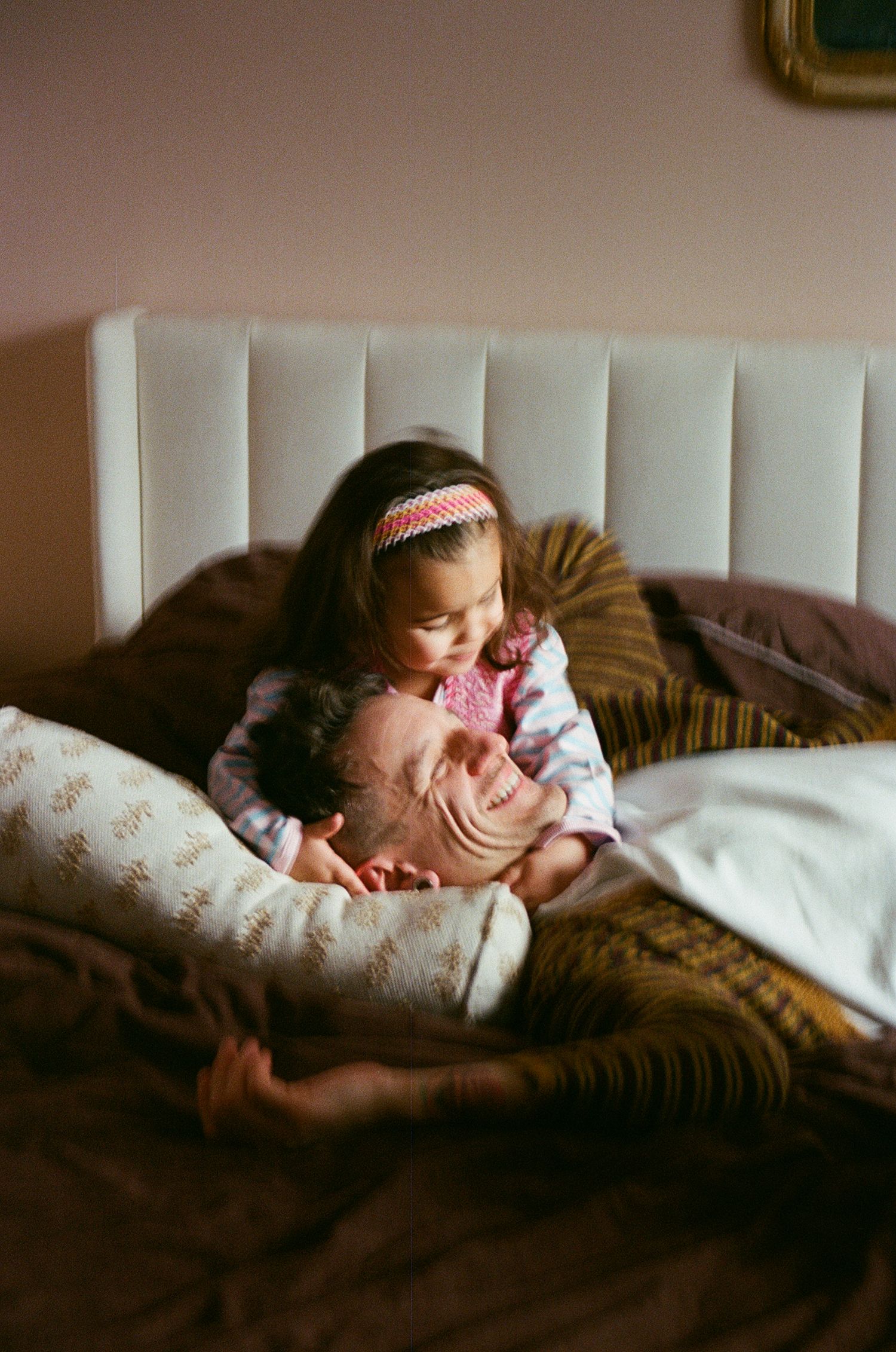 Young girl with headband cuddles smiling man lying on brown bed with white pillow beside them.