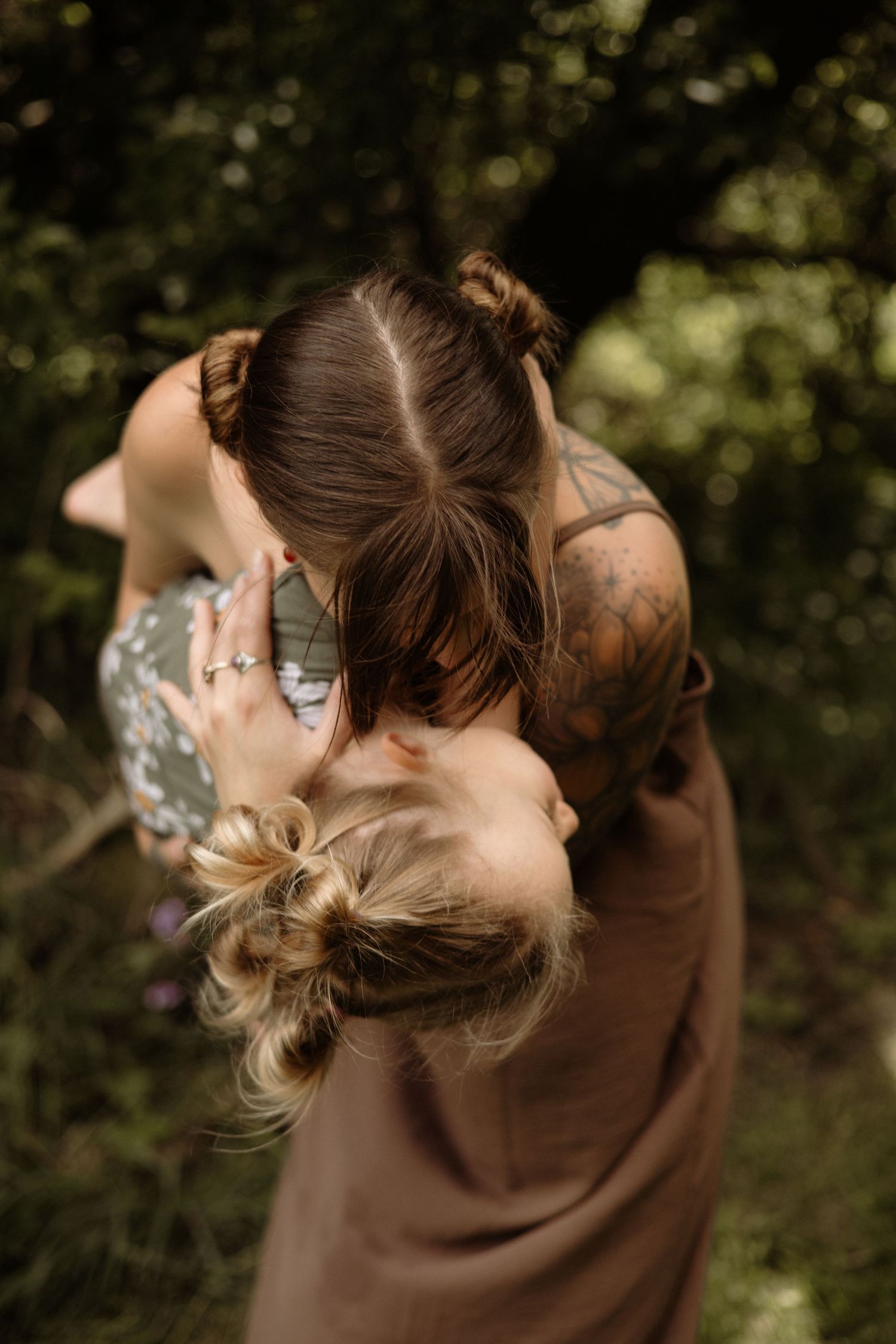 A tattooed mother embraces her young blonde daughter in a lush green outdoor setting.