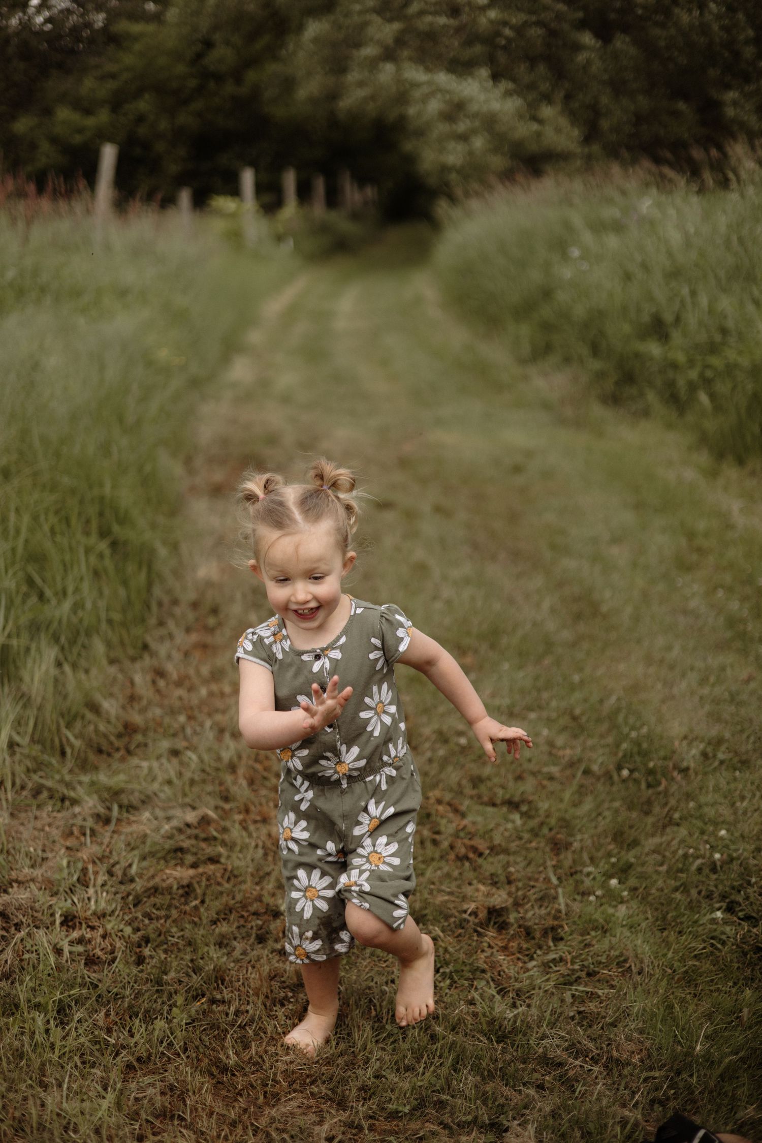 Laughing barefoot toddler girl in green daisy romper runs joyfully along grassy outdoor path.