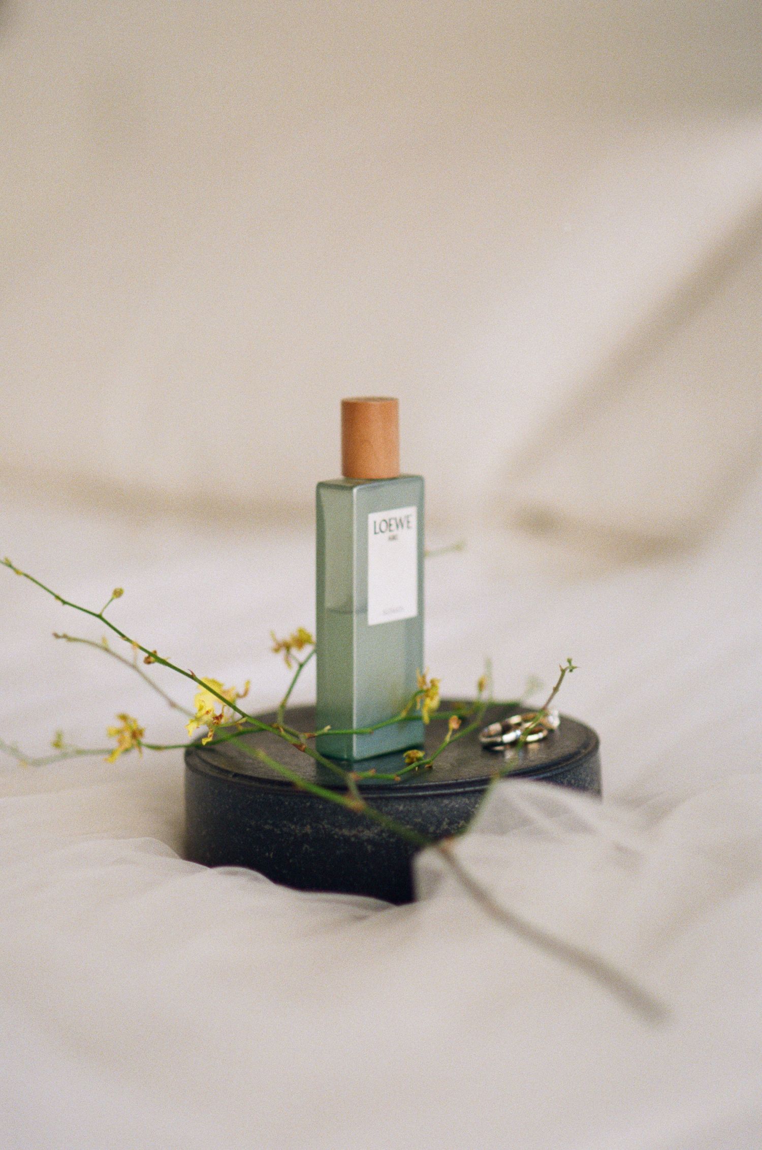 Loewe green glass perfume bottle with cork cap displayed on a dark stand with yellow wildflowers.