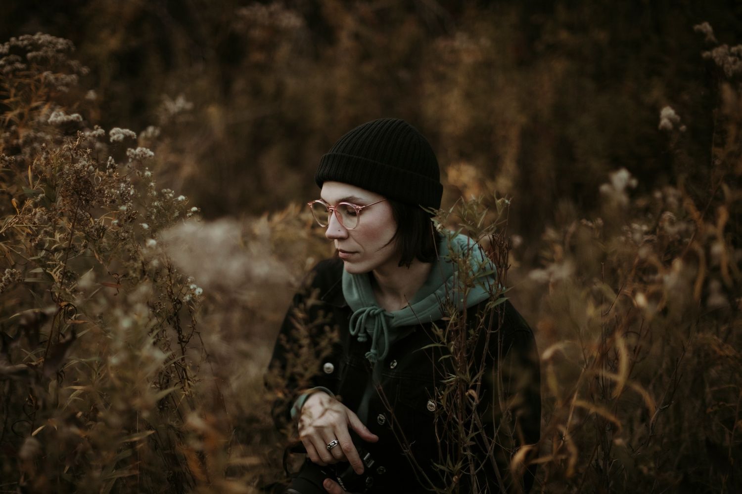 Woman in black beanie sitting among dried autumn wildflowers in a moody outdoor portrait.