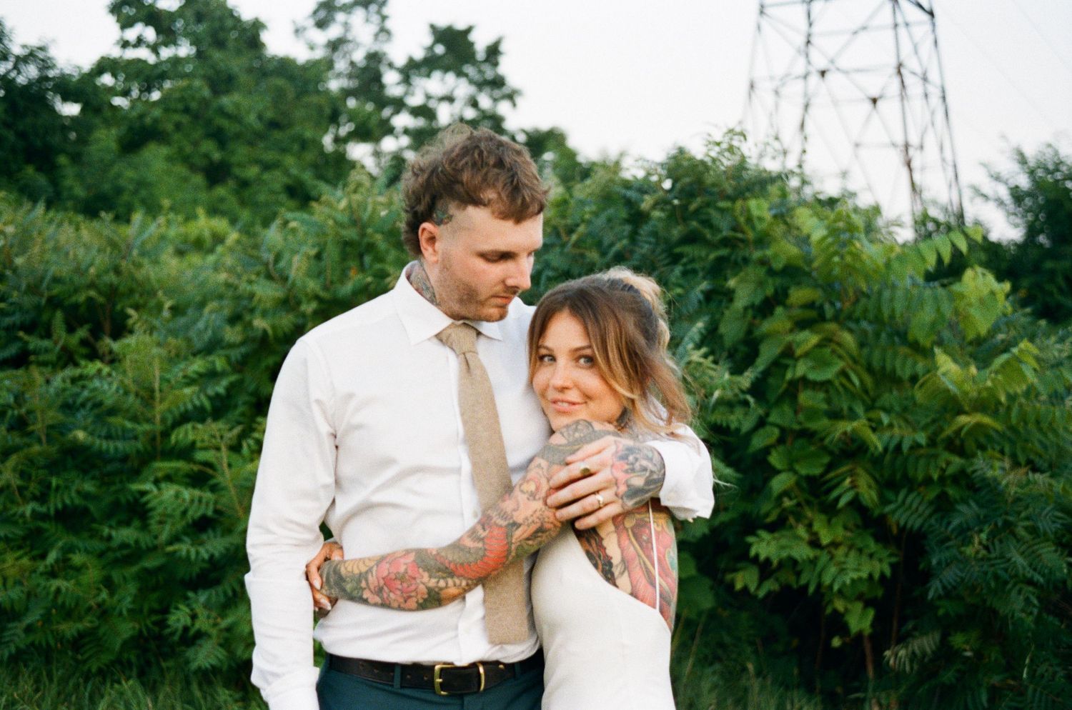 Tattooed bride and groom embracing in a lush green outdoor setting on their wedding day.