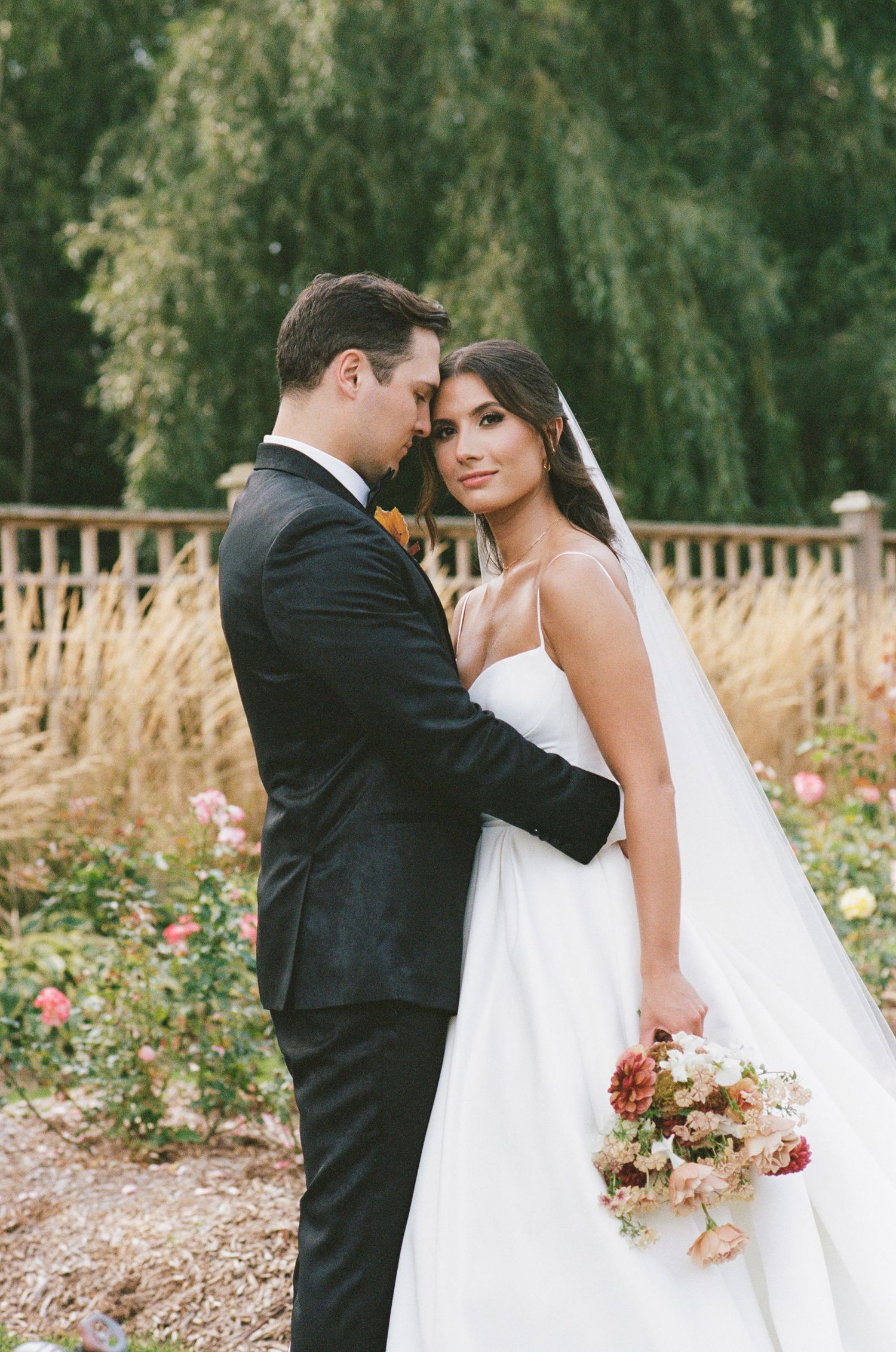 Bride and groom in an intimate embrace in a romantic garden setting with willow trees in background.
